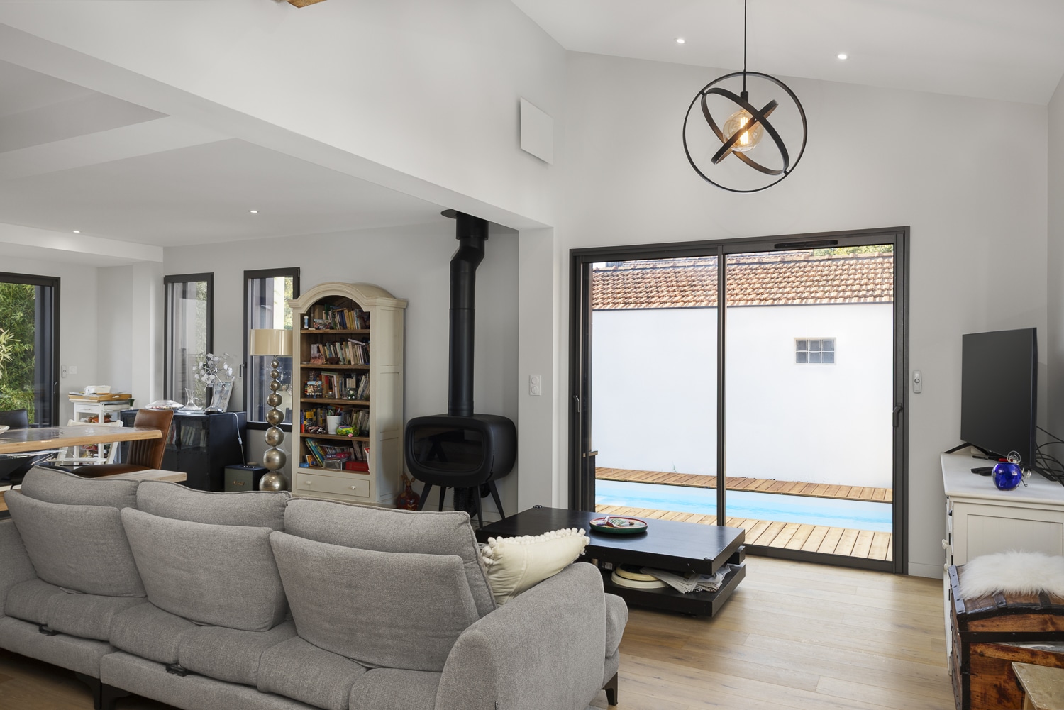 Salon moderne avec canapé gris, poêle à bois noir, bibliothèque crème et baie vitrée donnant sur une piscine extérieure.