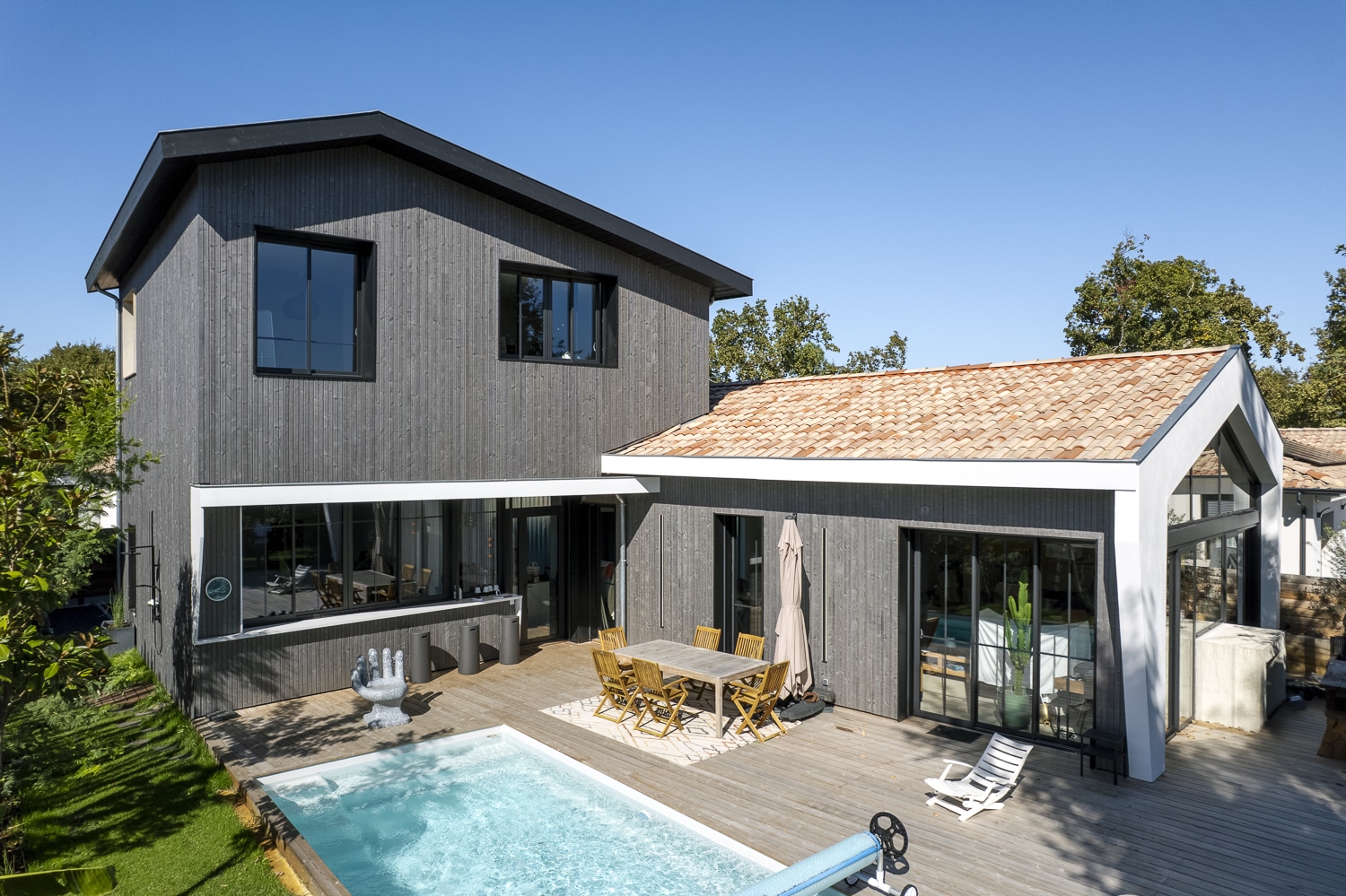 Maison moderne bardage bois gris avec piscine extérieure, terrasse bois et une annexe au toit de tuiles rouges sous un ciel bleu.