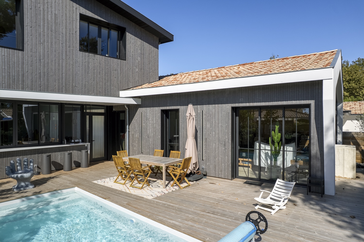 Moderne maison bois gris avec terrasse bois, piscine extérieure, table à manger, chaise main sculptée et parasol beige.
