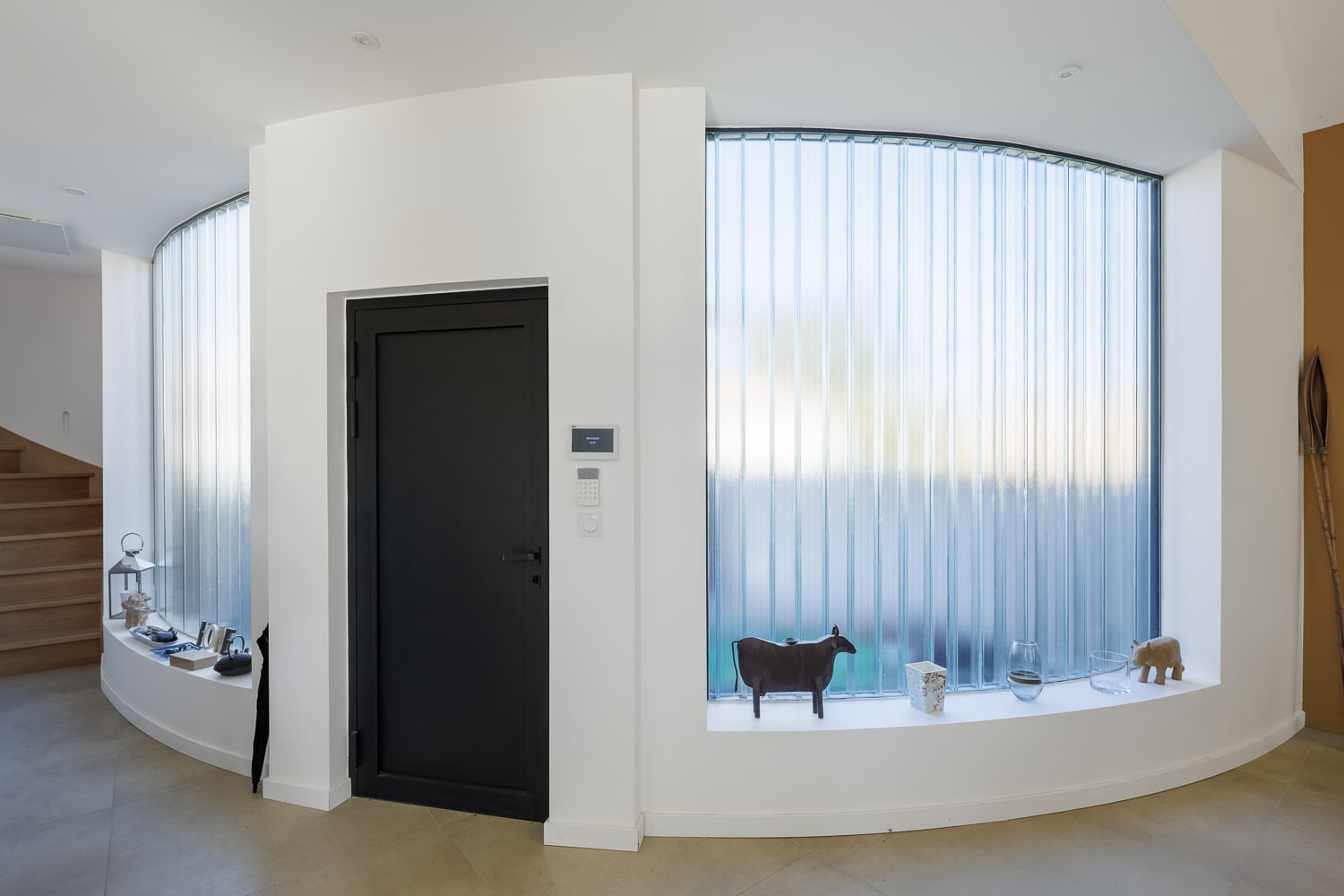 Hall d'entrée moderne avec mur blanc, porte noire, et grande fenêtre incurvée en verre nervuré laissant filtrer la lumière.