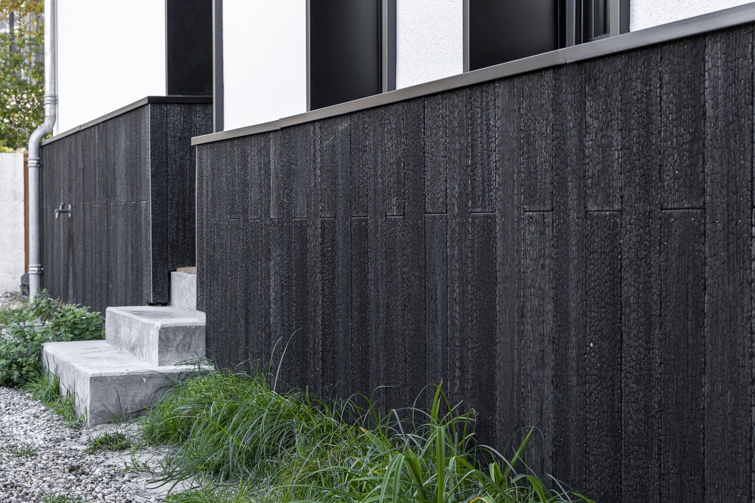 Bardage bois brûlé noirci (Shou Sugi Ban) d'une façade moderne avec escalier en béton et herbes vertes.