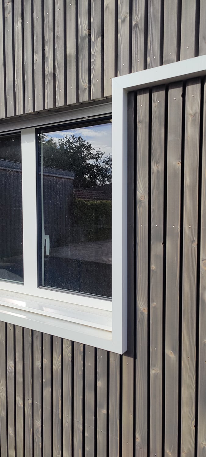 Détail d'une fenêtre blanche sur bardage vertical en bois teinté gris foncé, reflets d'arbres et ciel bleu sur la vitre.