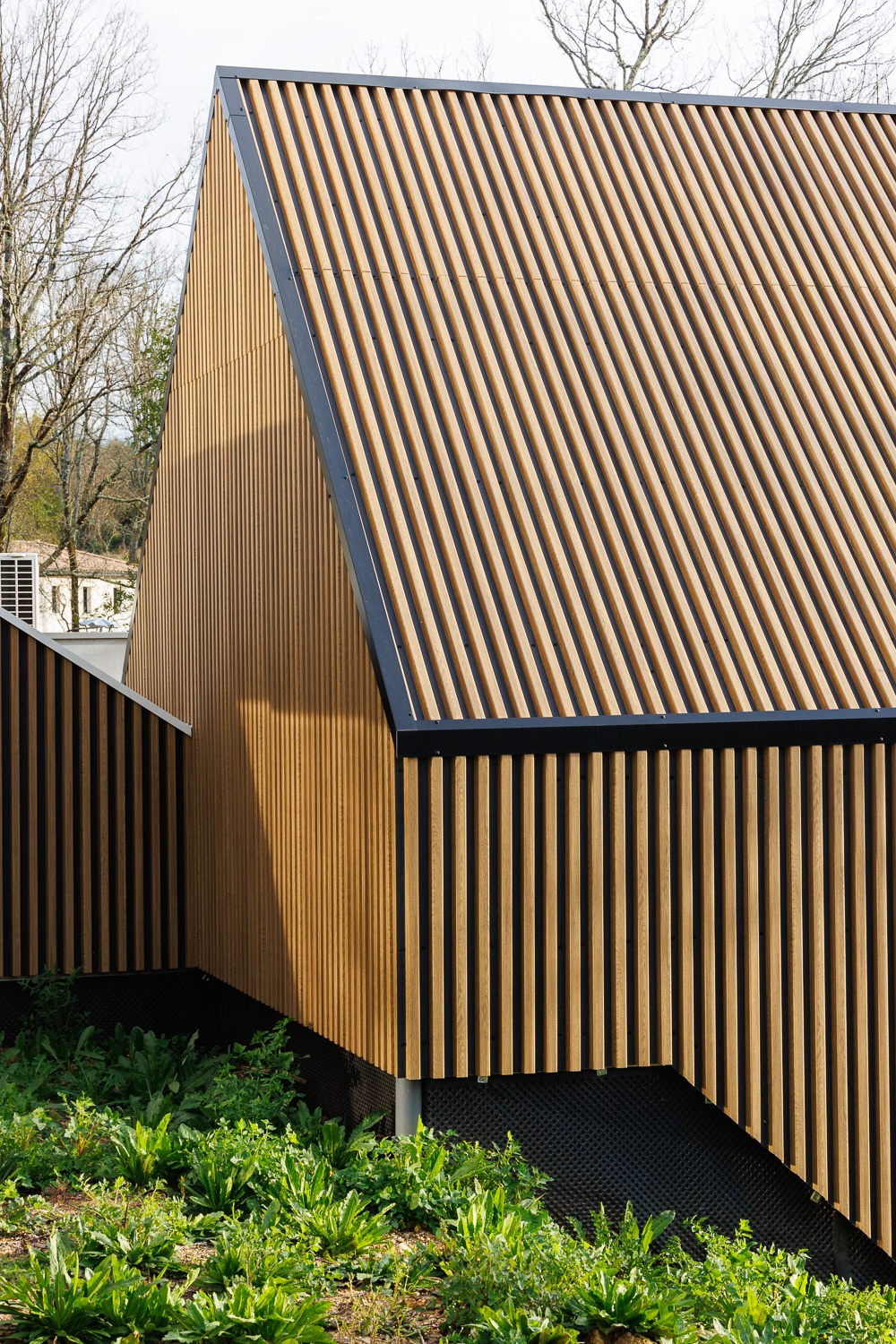 Bâtiment moderne avec bardage en lattes de bois verticales et toit en pente, entouré de verdure.