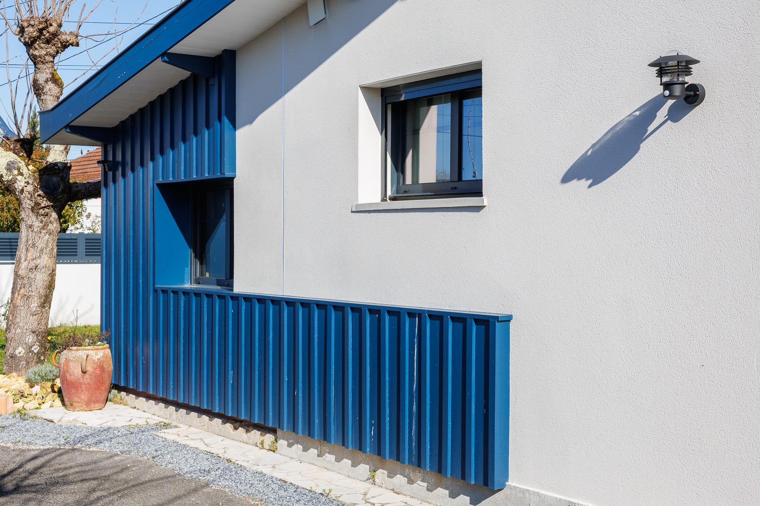 Bardage vertical bleu sur façade blanche de maison moderne avec luminaire noir et pot en terre cuite.