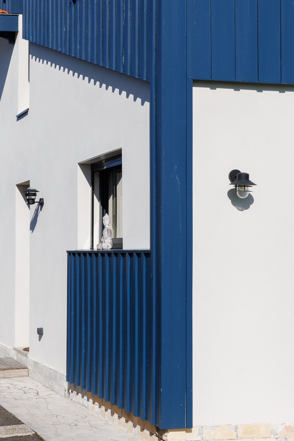 Maison moderne avec bardage bois bleu, façade blanche, statuette de Bouddha et luminaires extérieurs noirs.