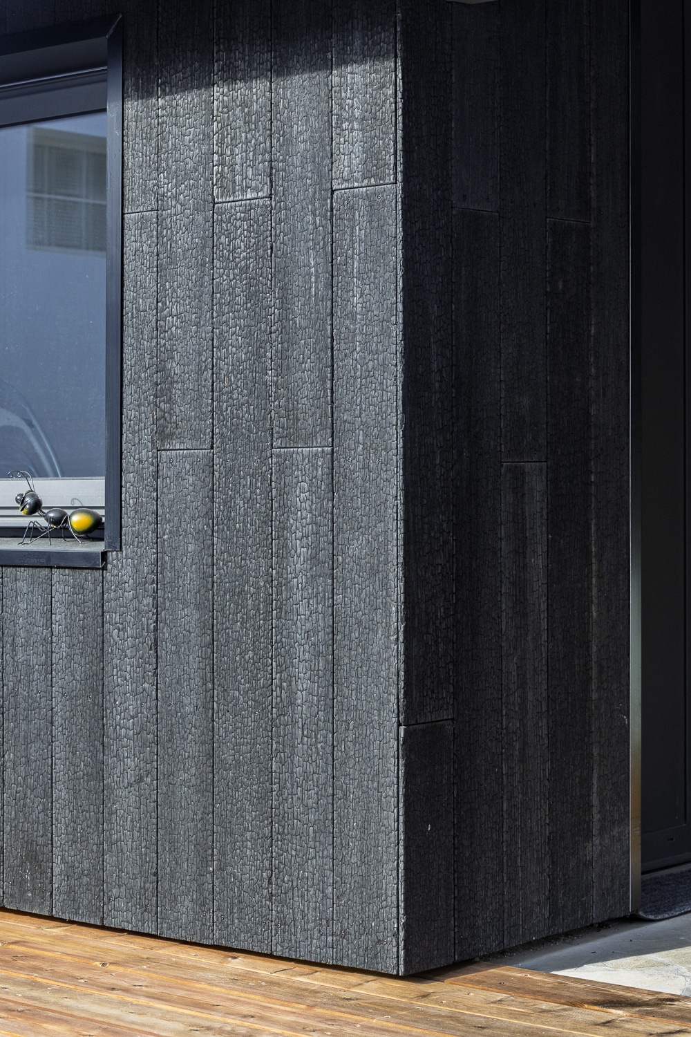 Muret extérieur à bardage bois brûlé Shou Sugi Ban noir, avec une sauterelle décorative jaune sur le rebord d'une fenêtre.