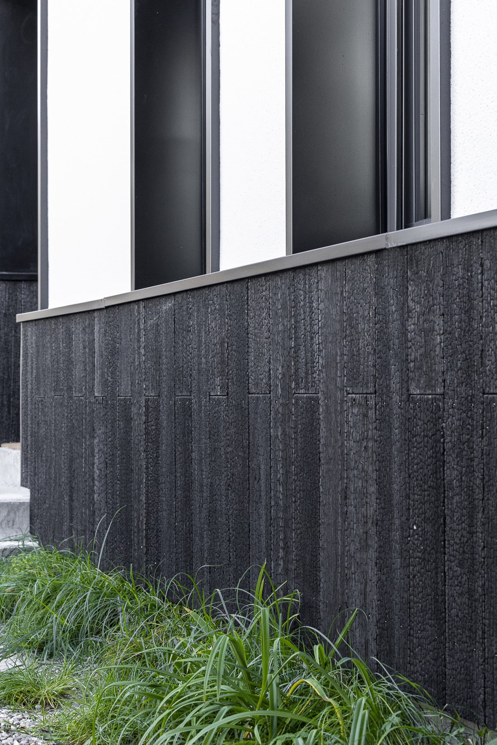 Bardage bois brûlé Shou Sugi Ban noir devant une façade blanche et des fenêtres sombres, avec de l'herbe verte au premier plan.