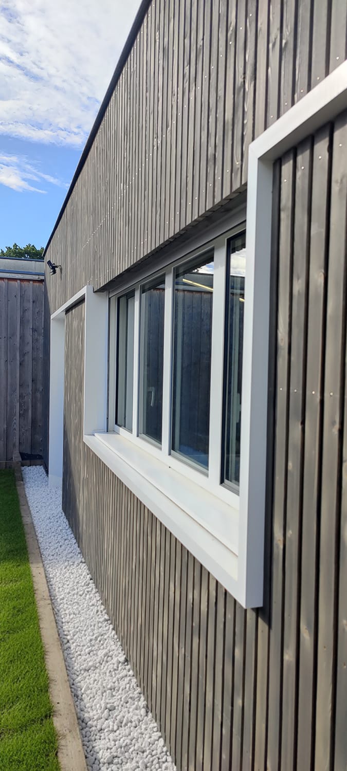 Façade d'un bâtiment moderne avec bardage bois gris foncé, grandes fenêtres blanches, et un chemin de gravier blanc longeant la pelouse.