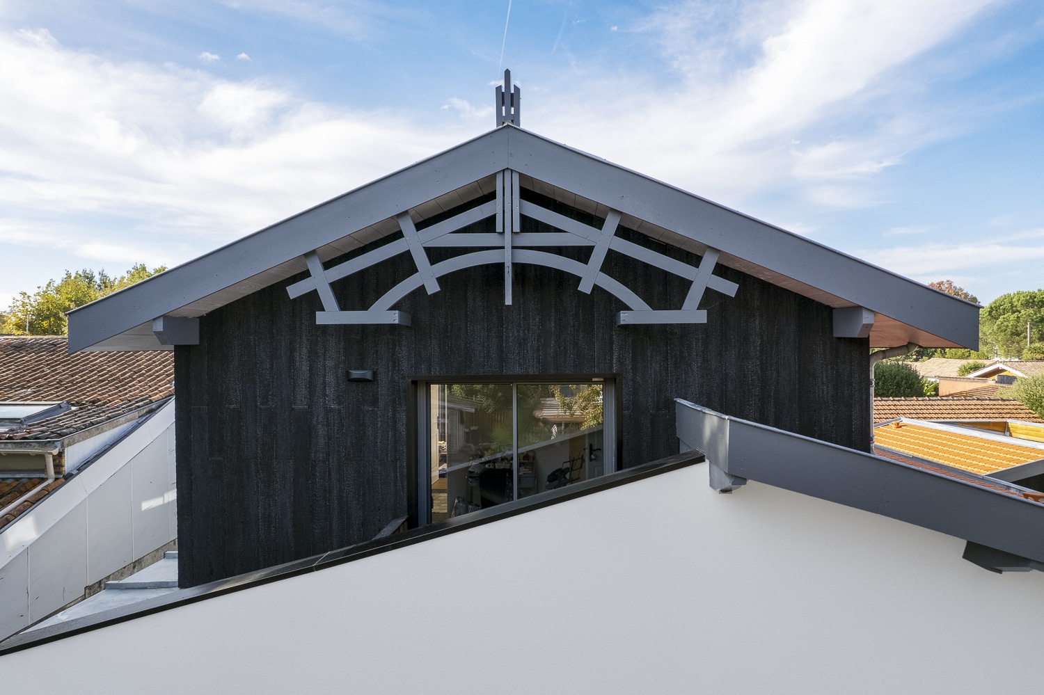 Dortoir d'inspiration basque ou chalet moderne avec bardage bois brûlé noir, pignon orné de fermes décoratives grises sous un ciel bleu.