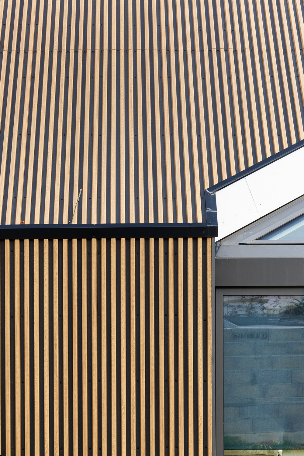 Bardage vertical en tasseaux de bois clair sur façade et toiture d'une maison moderne avec menuiserie grise.