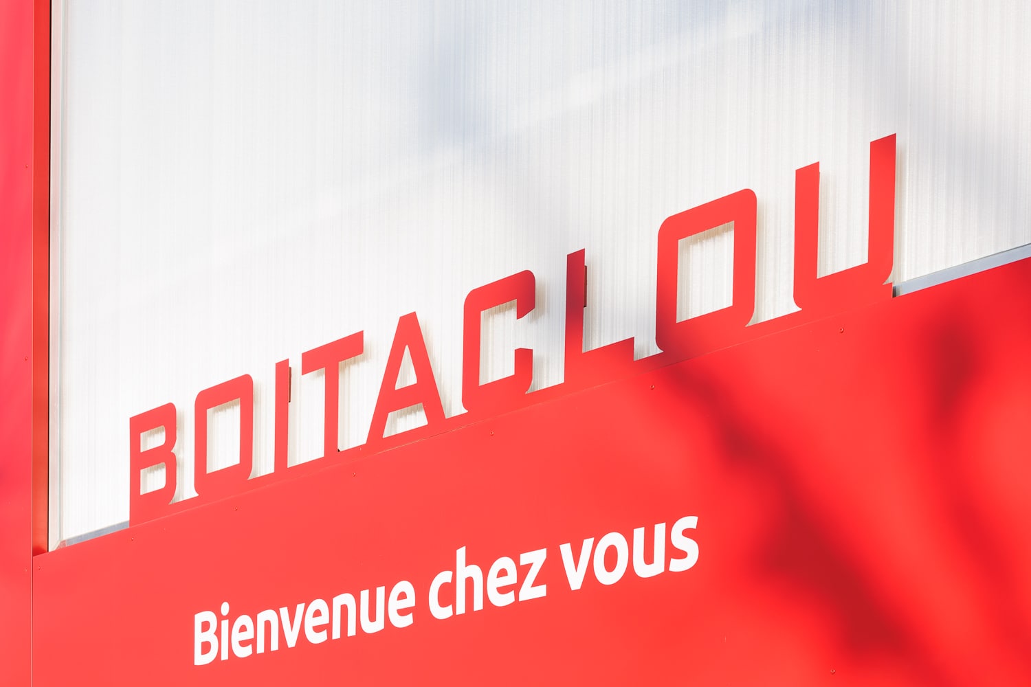 Enseigne rouge Boitaclou avec le slogan Bienvenue chez vous sur un fond blanc texturé moderne et accueillant.