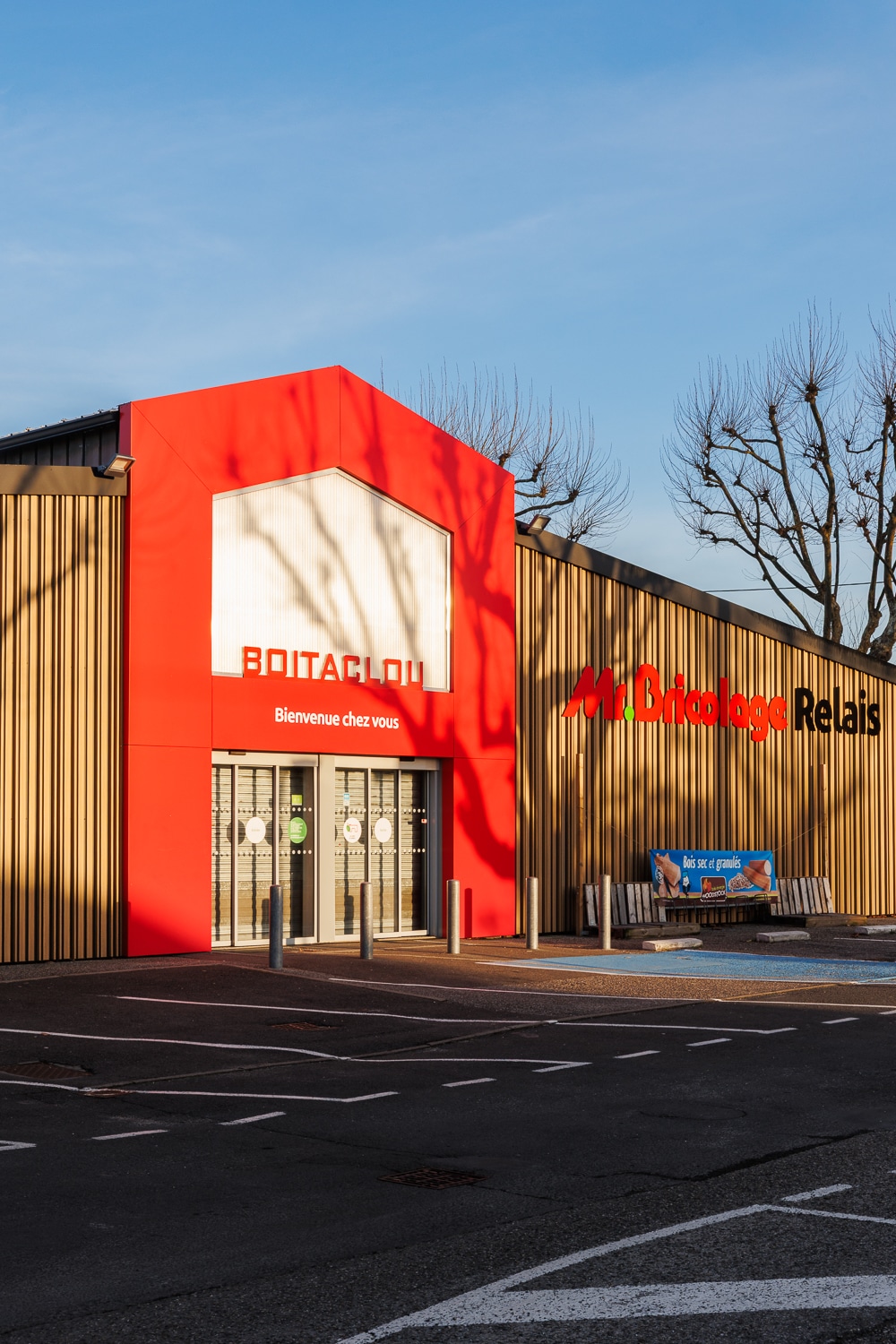 Entrée rouge du magasin Boitaclou et Mr.Bricolage Relais sous un ciel bleu dégagé. Bienvenue chez vous.