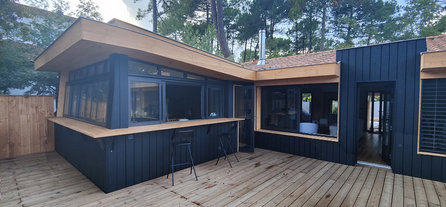 Cabane de jardin moderne en bois noir et clair avec bar extérieur et terrasse, sous des pins. Toit tuiles et cheminée.
