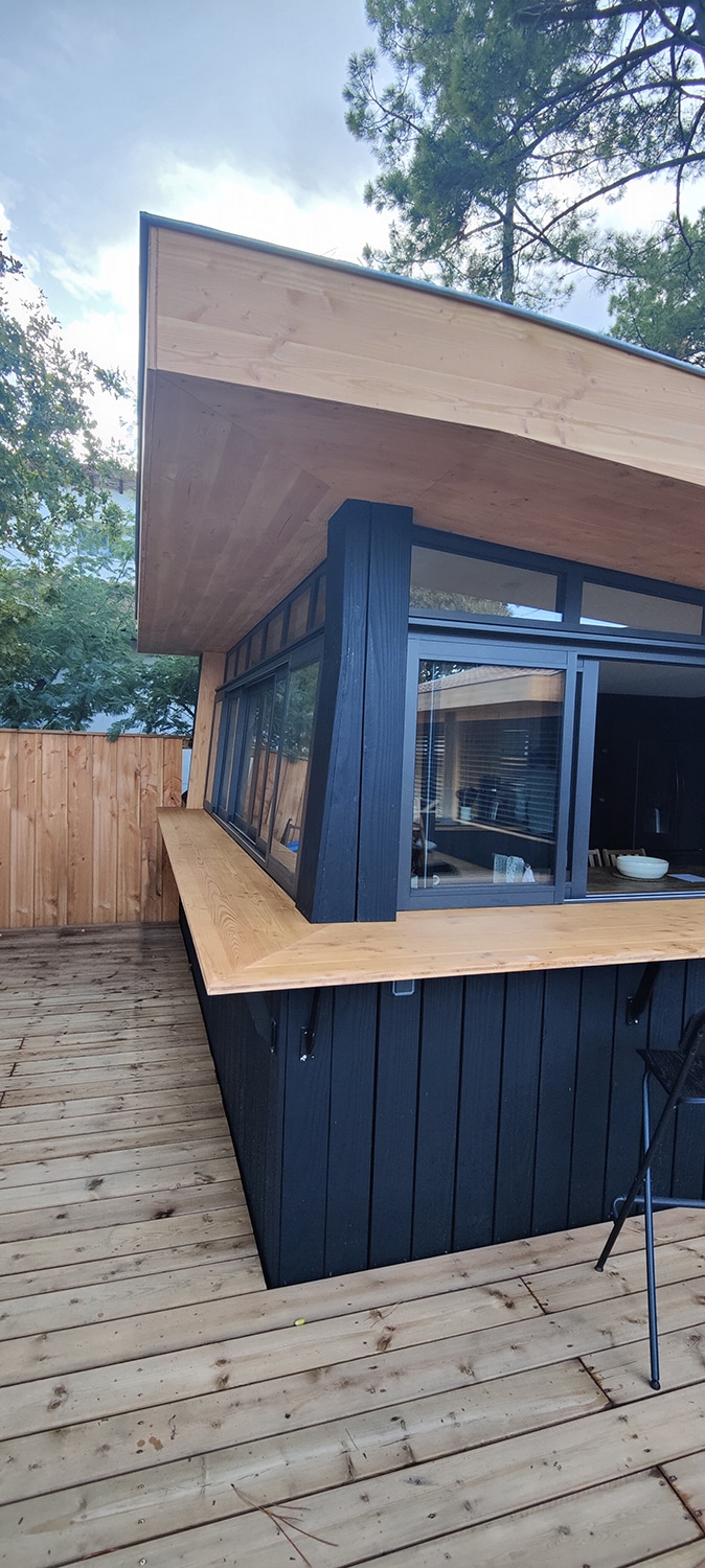 Structure extérieure moderne en bois clair et noir sur terrasse, grand comptoir et fenêtres, ciel nuageux, arbres.