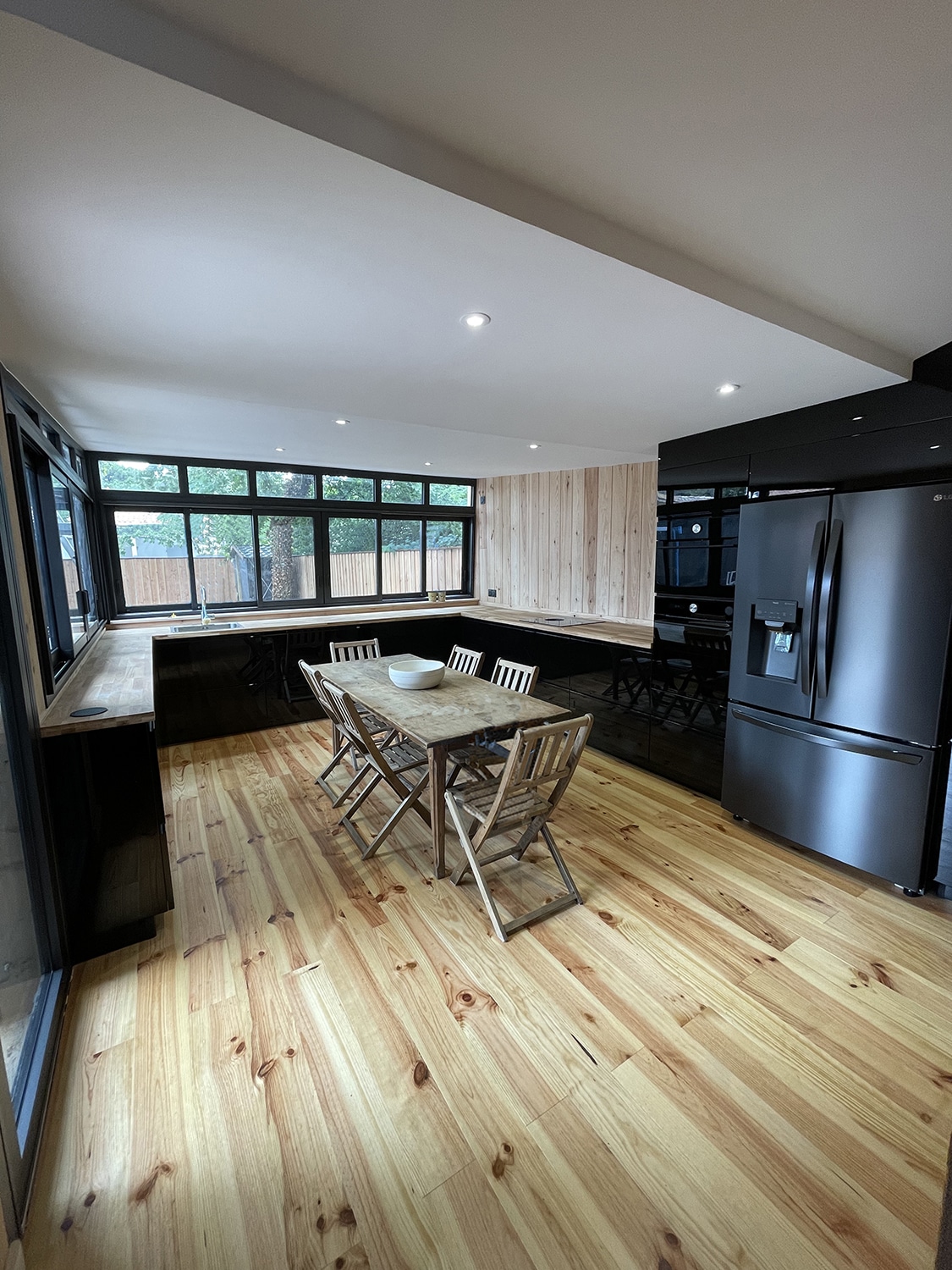Cuisine ouverte moderne avec placards noirs, comptoirs et plancher en bois clair, réfrigérateur LG, table rustique et larges fenêtres.