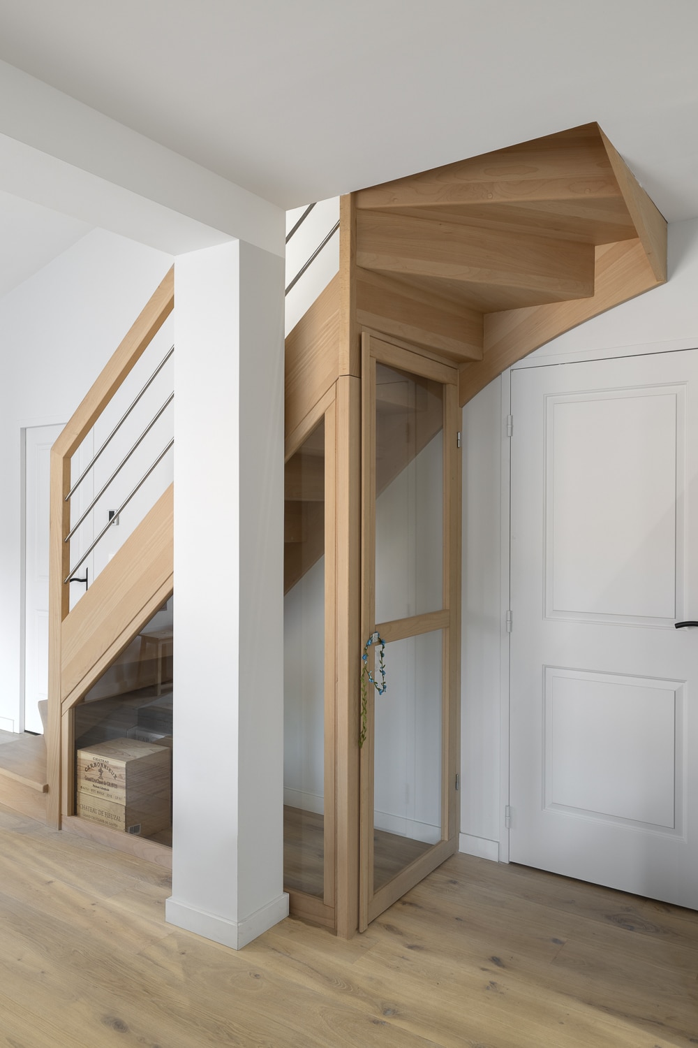 Escalier en bois clair avec rampe métallique et ascenseur vitré encastré, près d'une colonne blanche et porte blanche.