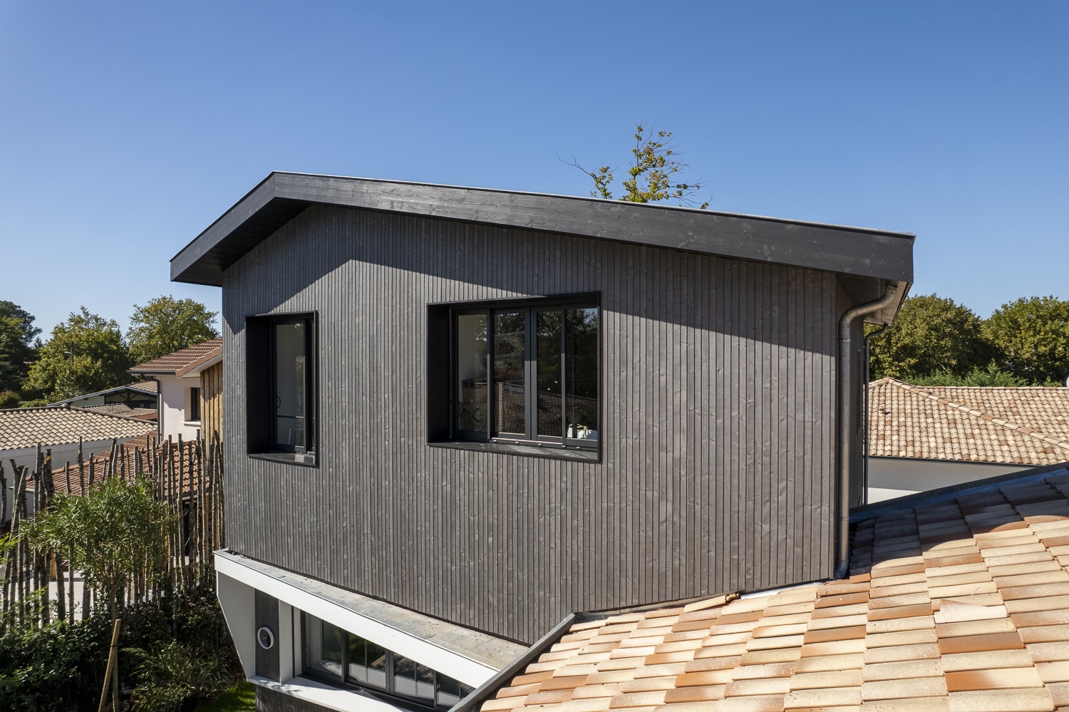 Agencement moderne d'une extension de maison avec bardage bois gris foncé vertical et toiture inclinée, vue sur les tuiles terre cuite voisines.
