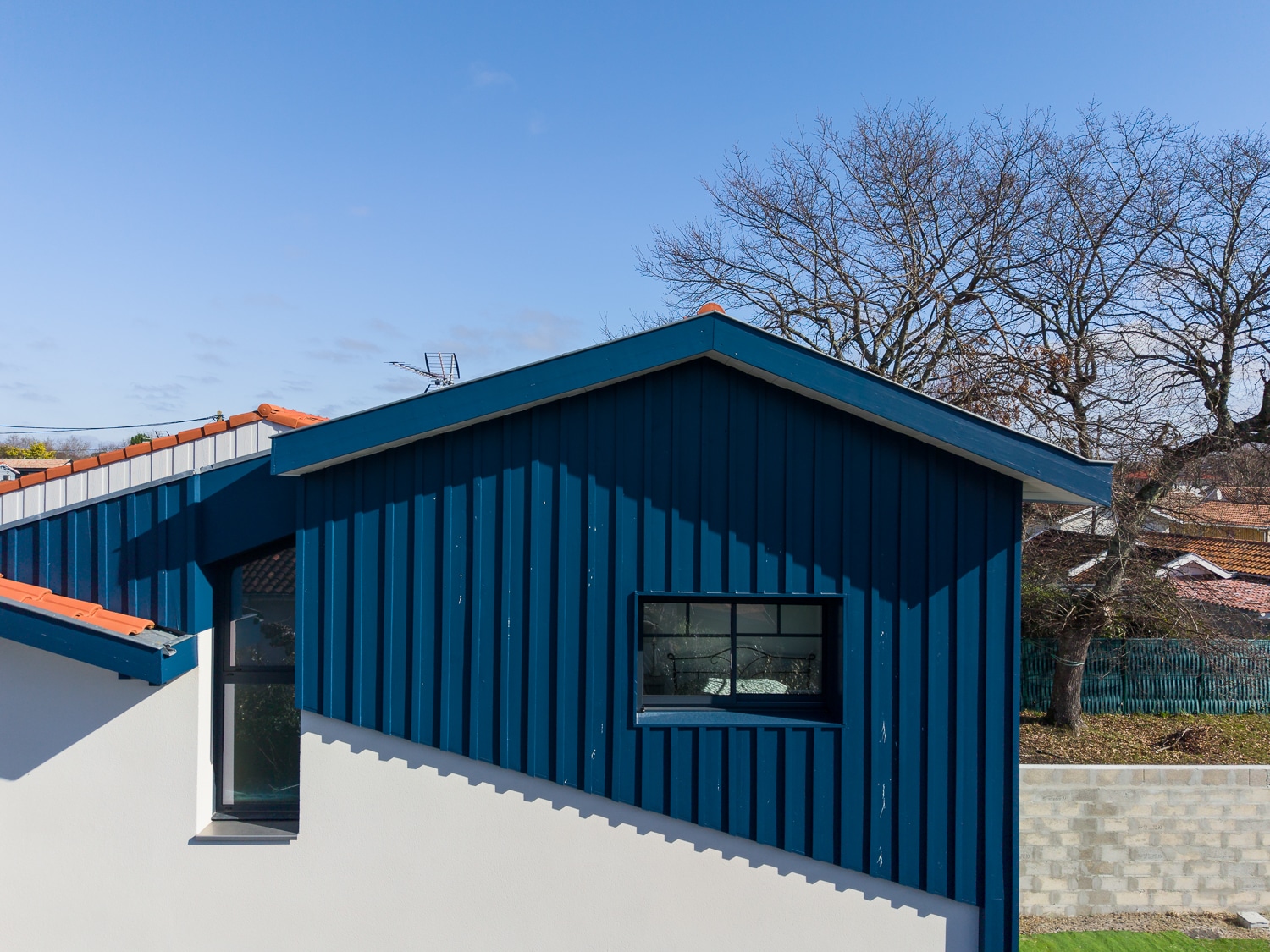 Extension de maison moderne avec bardage bois bleu canard sur façade blanche et toit en pente.