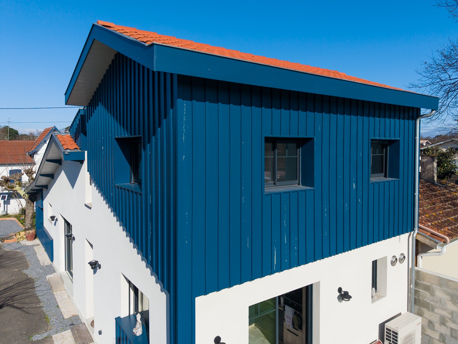 Extension de maison moderne avec bardage bois bleu vertical et façade blanche sous un ciel ensoleillé.