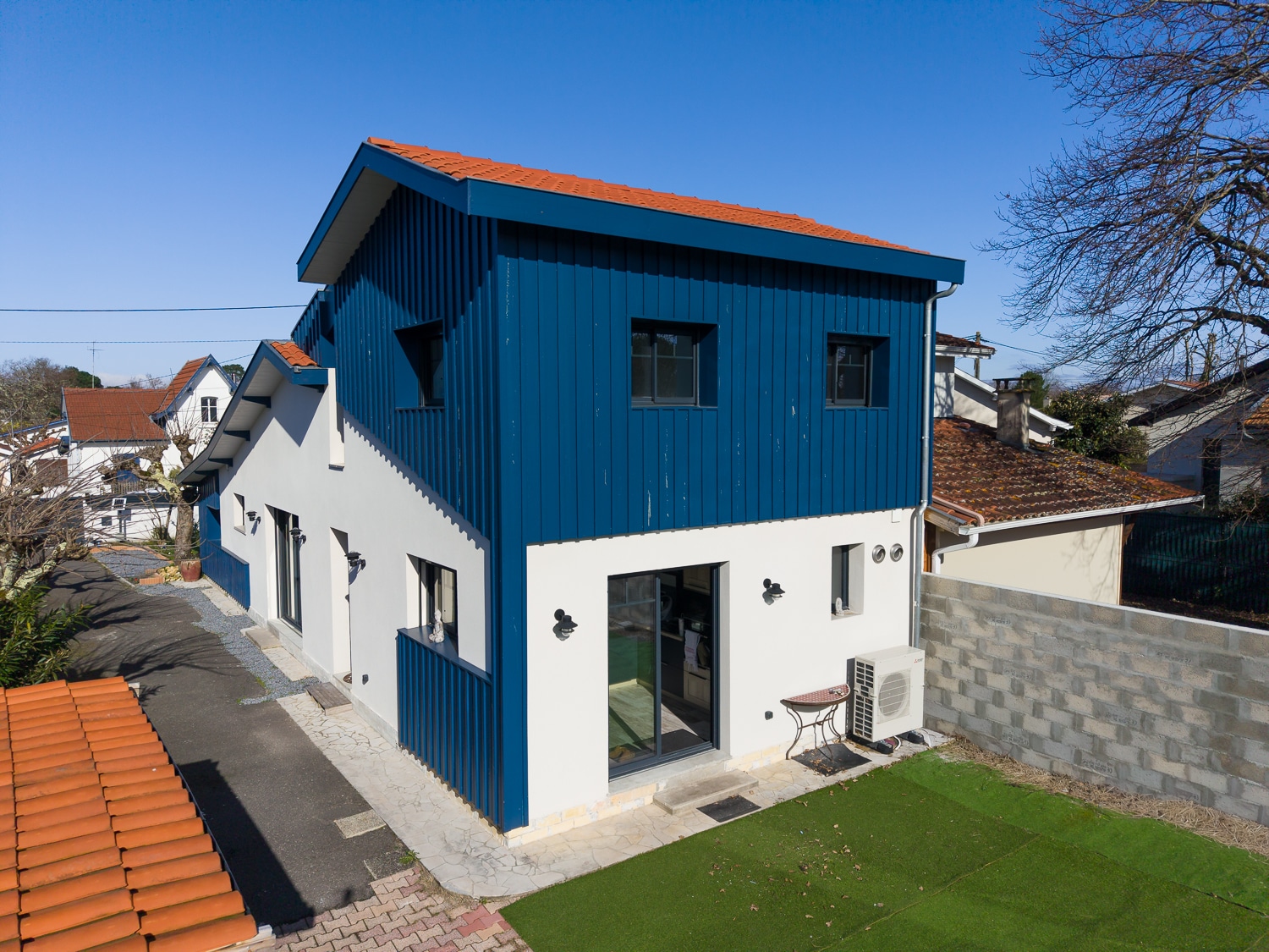 Maison moderne blanche avec extension à bardage vertical bleu, toit en tuiles rouges et pelouse synthétique.