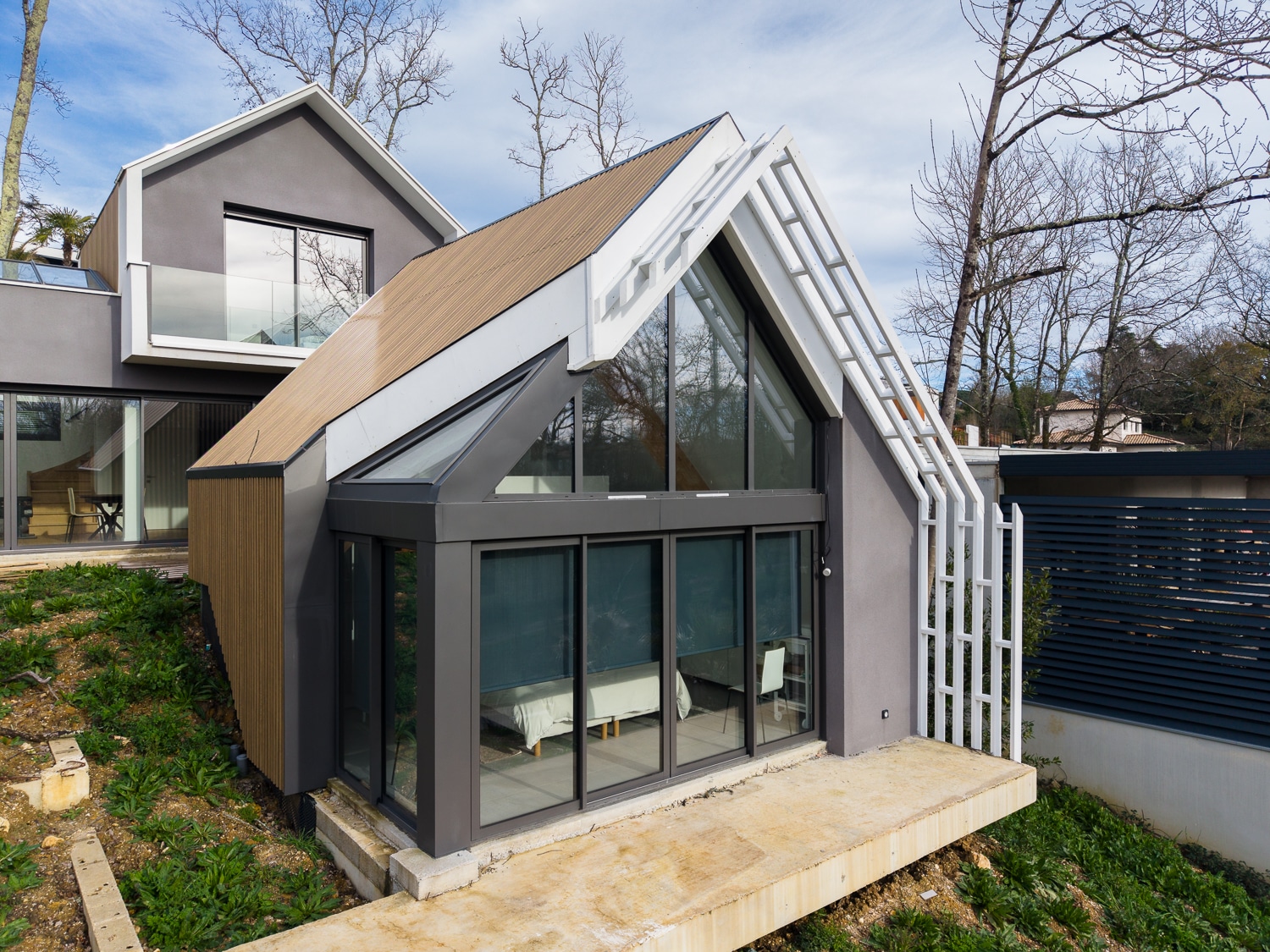 Extension de maison moderne avec toit incliné en bois et grandes baies vitrées sur un jardin en pente.
