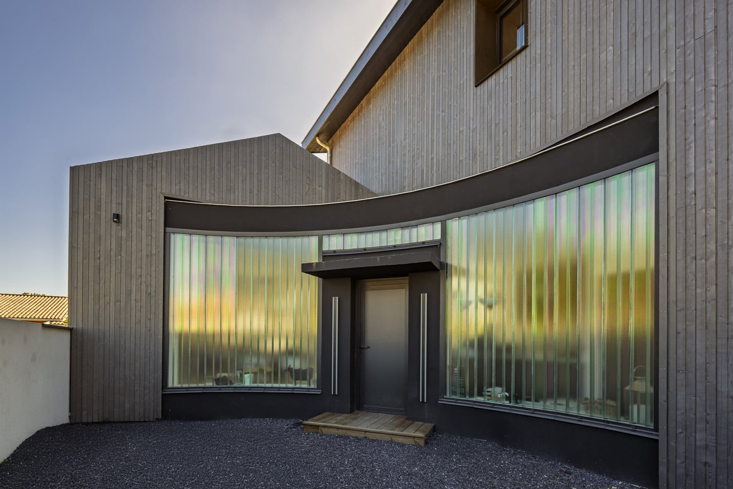 Façade moderne avec bardage bois foncé, entrée cintrée et grandes vitres verticales iridescentes captant la lumière.