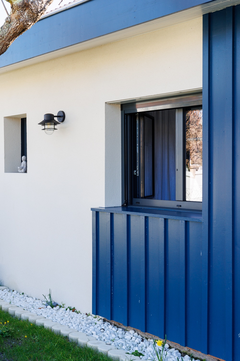 Façade moderne blanche avec bardage bois bleu, fenêtre noire, applique murale et parterre de galets blancs au jardin.