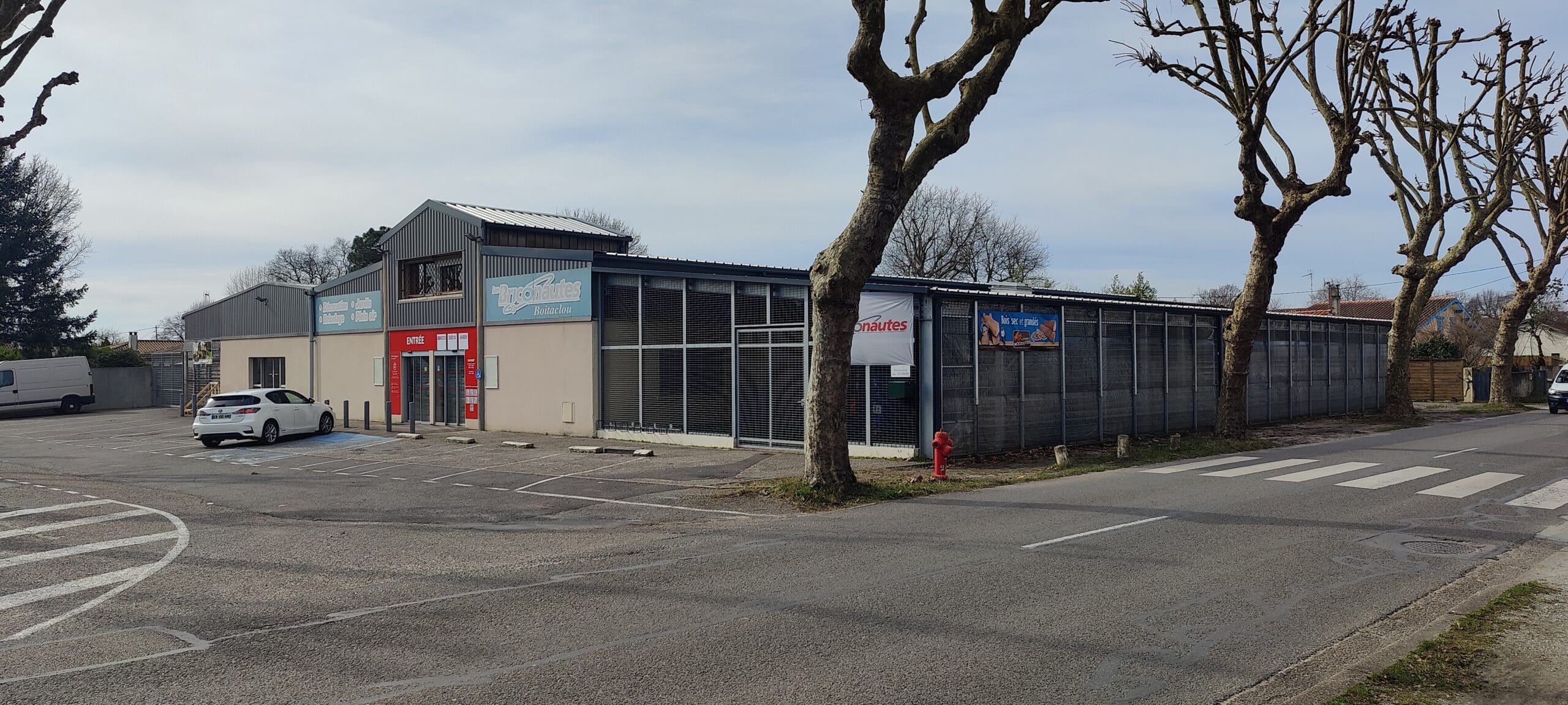 Devanture du magasin Les Briconautes Boitaclou, spécialisé en bricolage et jardin, avec une voiture blanche sur le parking.