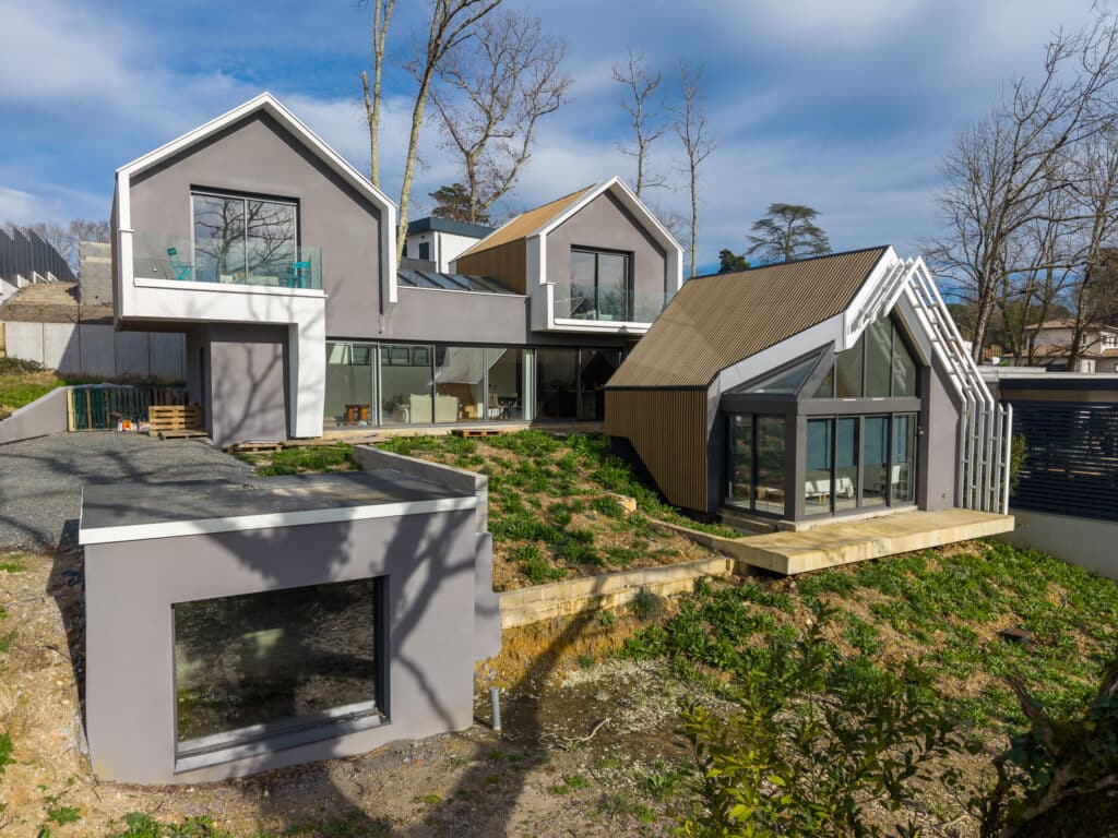 Maison contemporaine grise et bois avec toits à pignons et grandes baies vitrées sur un terrain paysager.