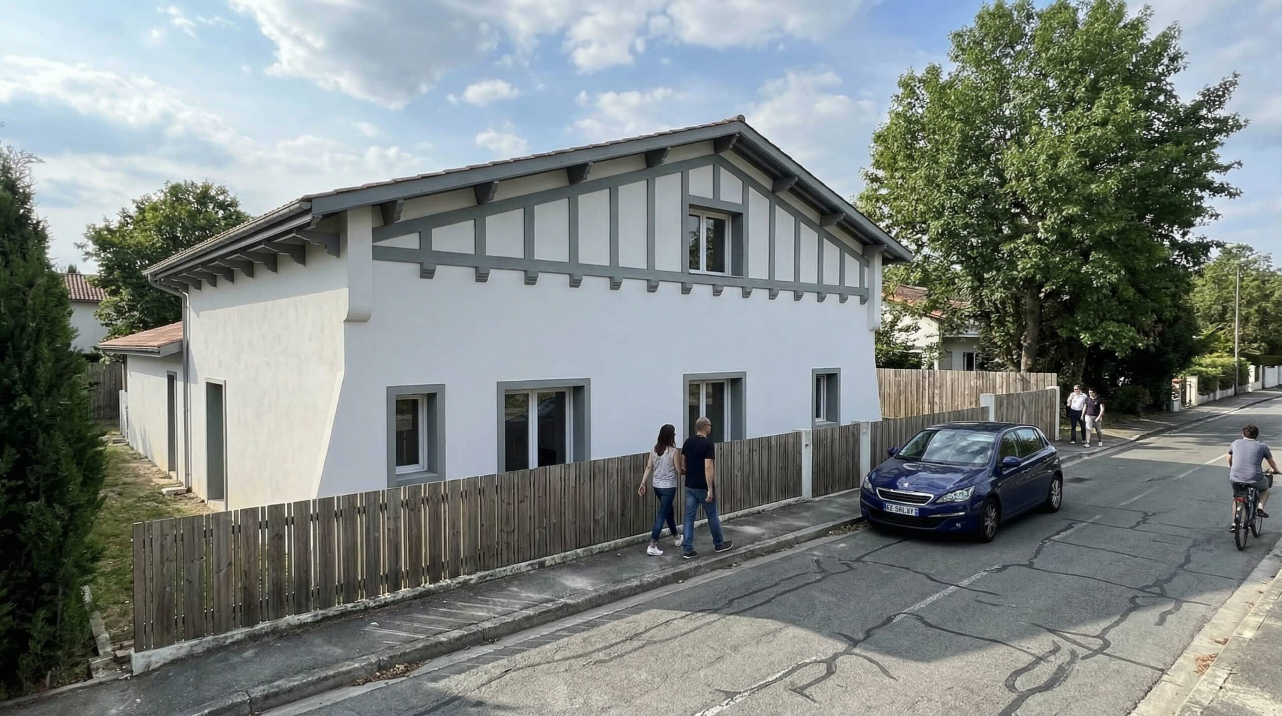 Maison basque blanche et grise, couple marchant sur le trottoir et Peugeot bleue garée dans une rue résidentielle.
