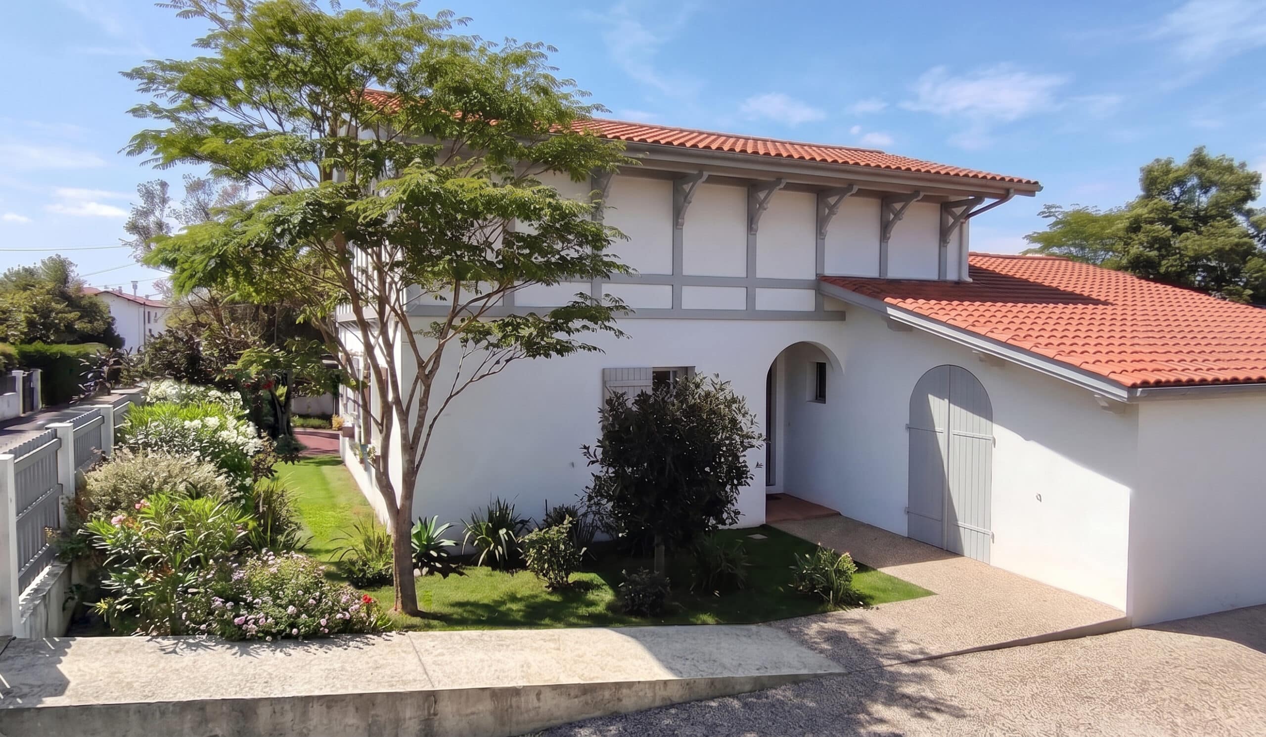 Maison basque blanche à toit de tuiles rouges avec jardin paysager et porte de garage grise sous un ciel bleu.