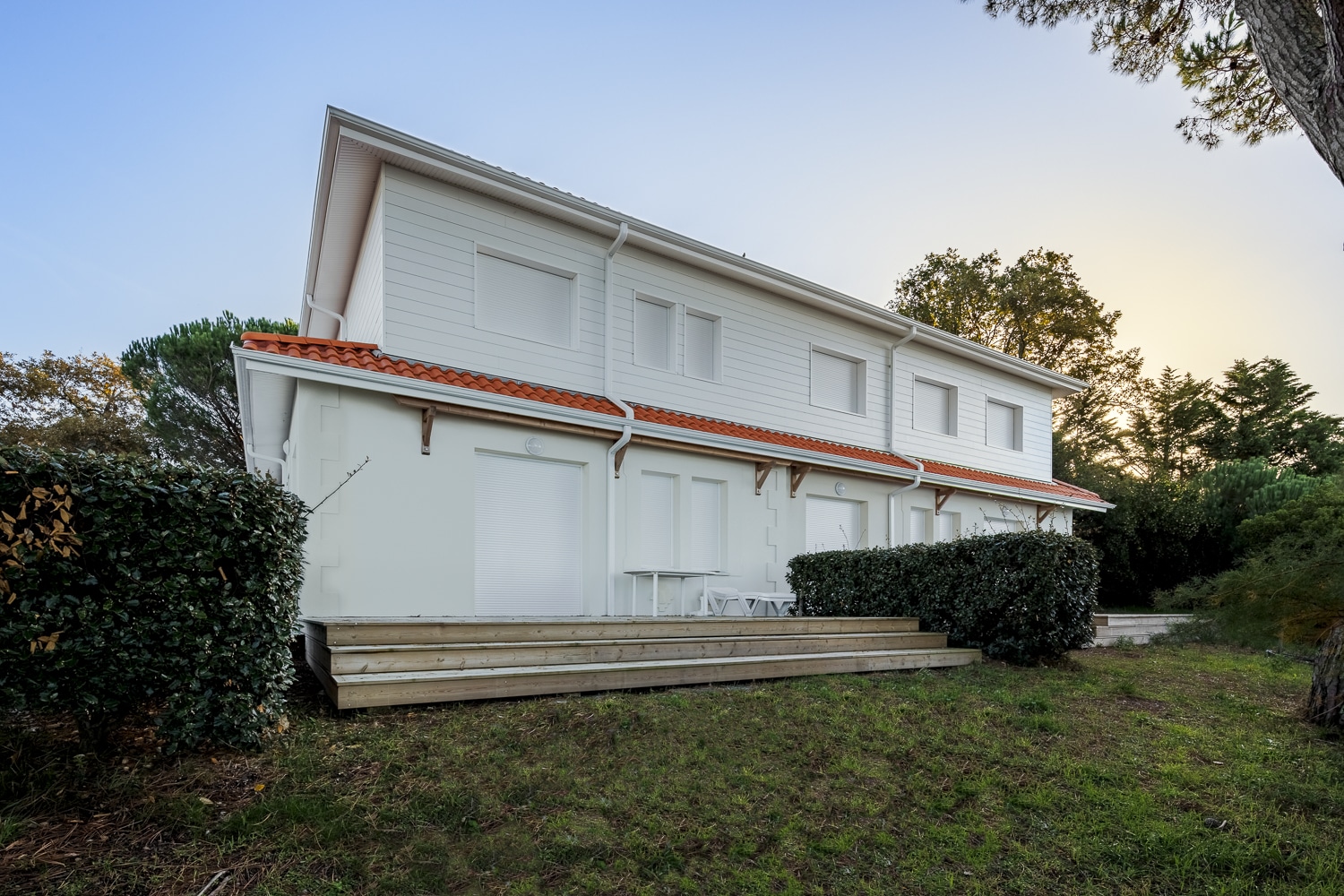 Longue maison blanche à deux étages avec toit de tuiles orange, terrasse en bois et haies vertes, sous un ciel bleu clair.