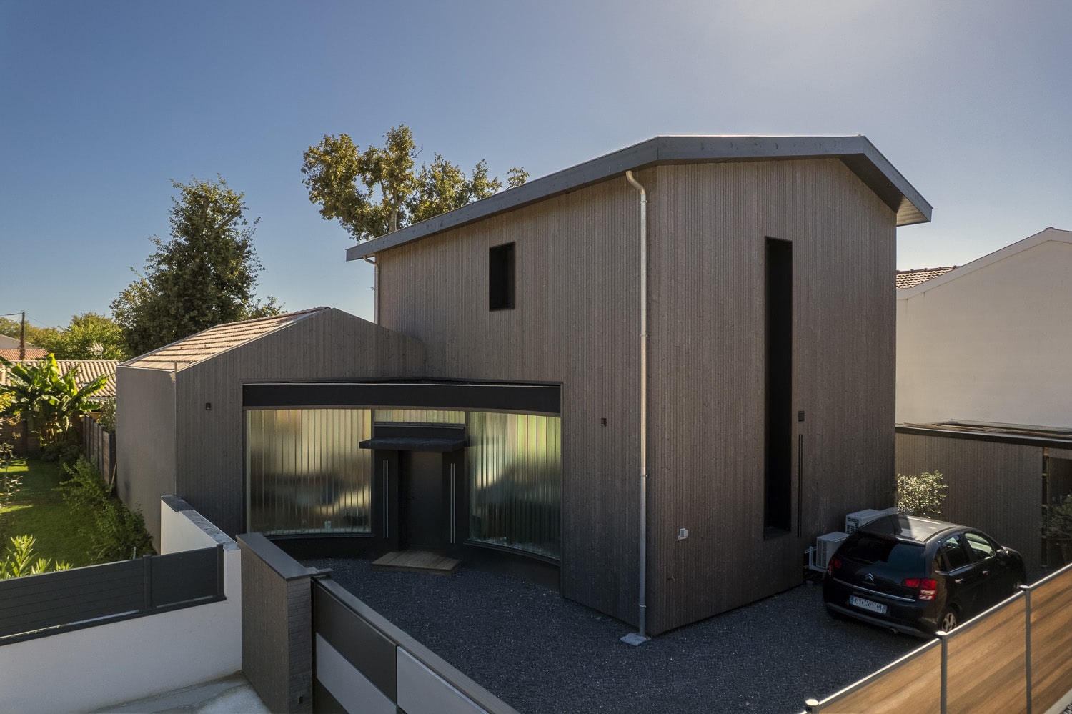 Maison moderne revêtue de bois brun foncé avec une entrée courbée et vitrée, sous un ciel bleu clair.