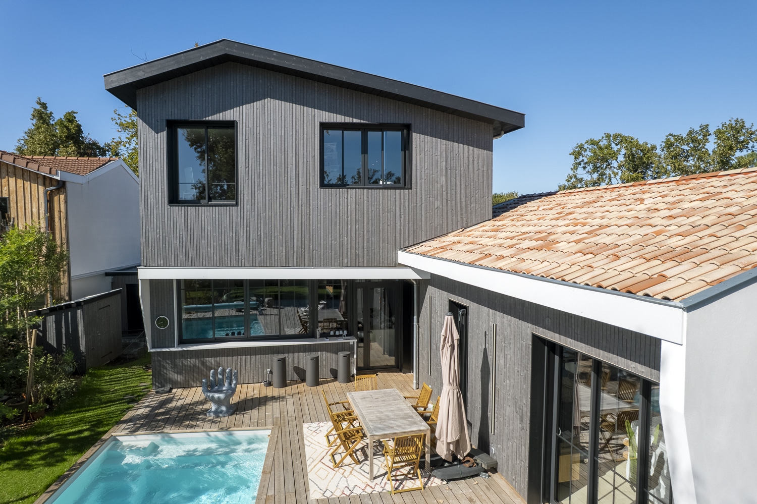 Maison moderne à bardage bois gris avec toit à tuiles rouges, terrasse en bois, piscine et sculpture de main.