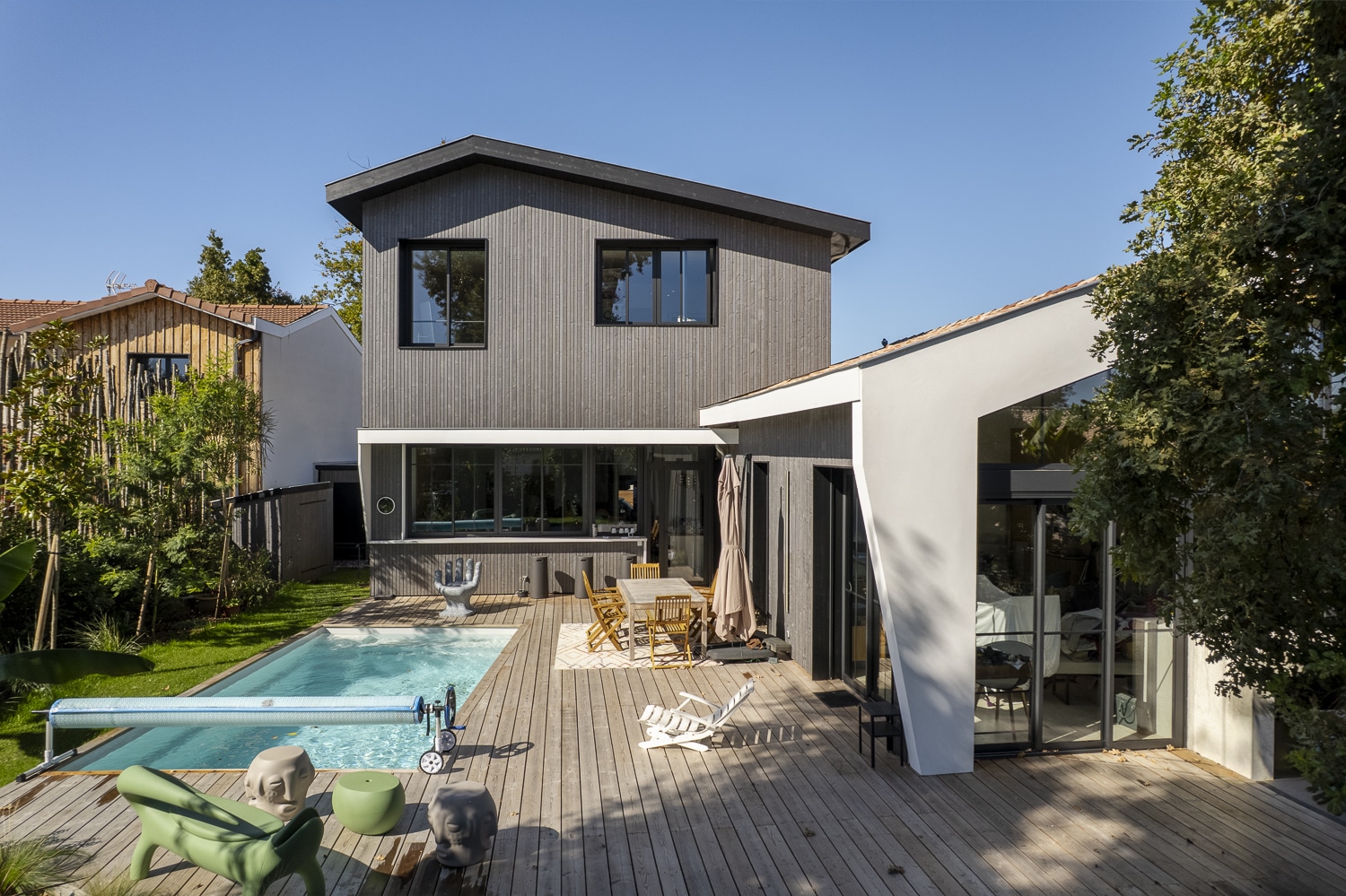 Terrasse en bois moderne avec piscine et mobilier design, juxtaposée à une maison à bardage gris et une extension blanche.