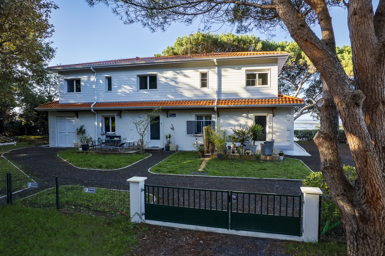 Maison à deux étages avec bardage clair et toit tuiles oranges, vue devant un portail vert et de grands arbres au premier plan. Panneau "P PARKING RESERVE".