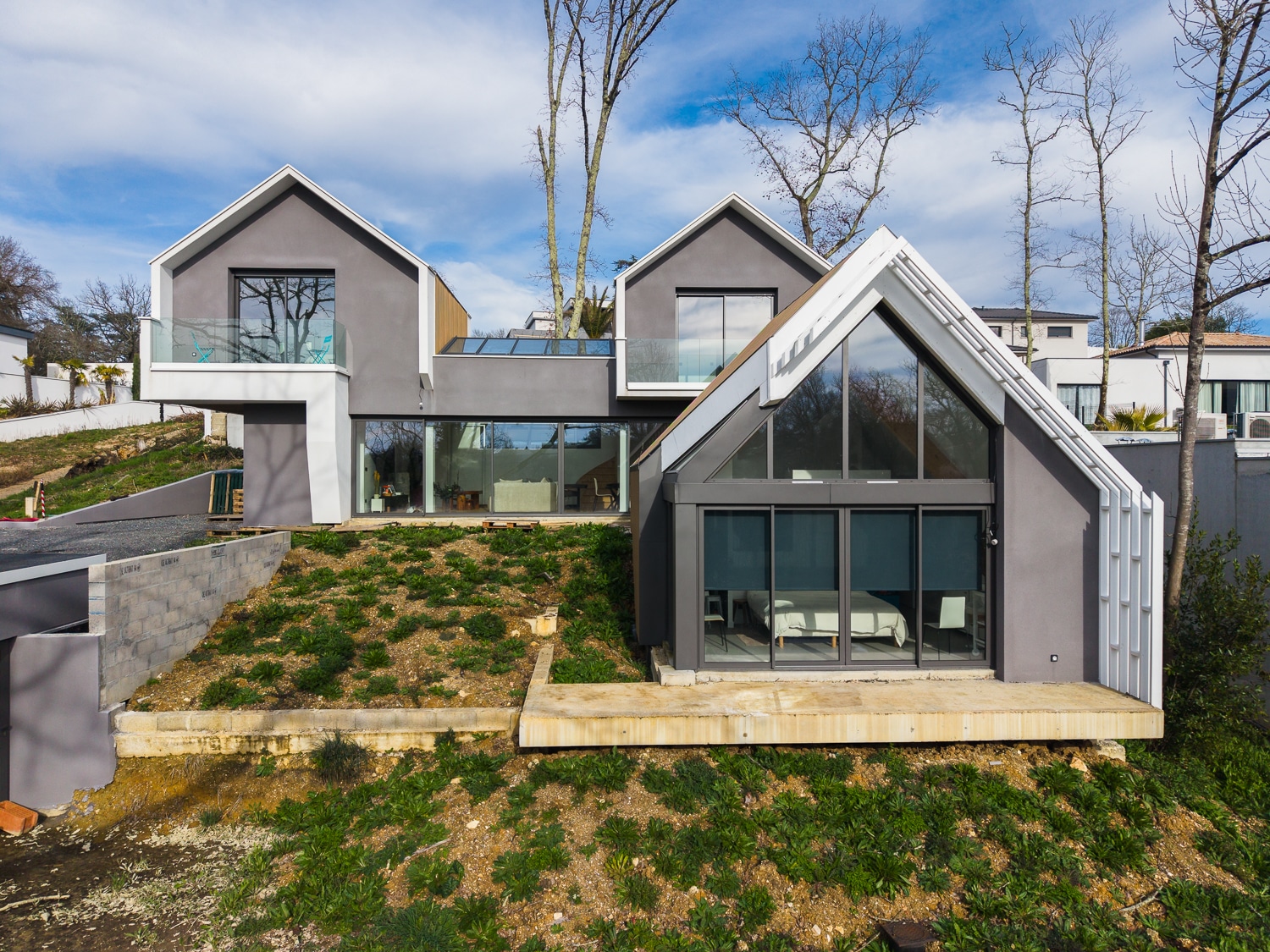 Maison contemporaine grise à toits pointus avec grandes baies vitrées sur terrain en pente.