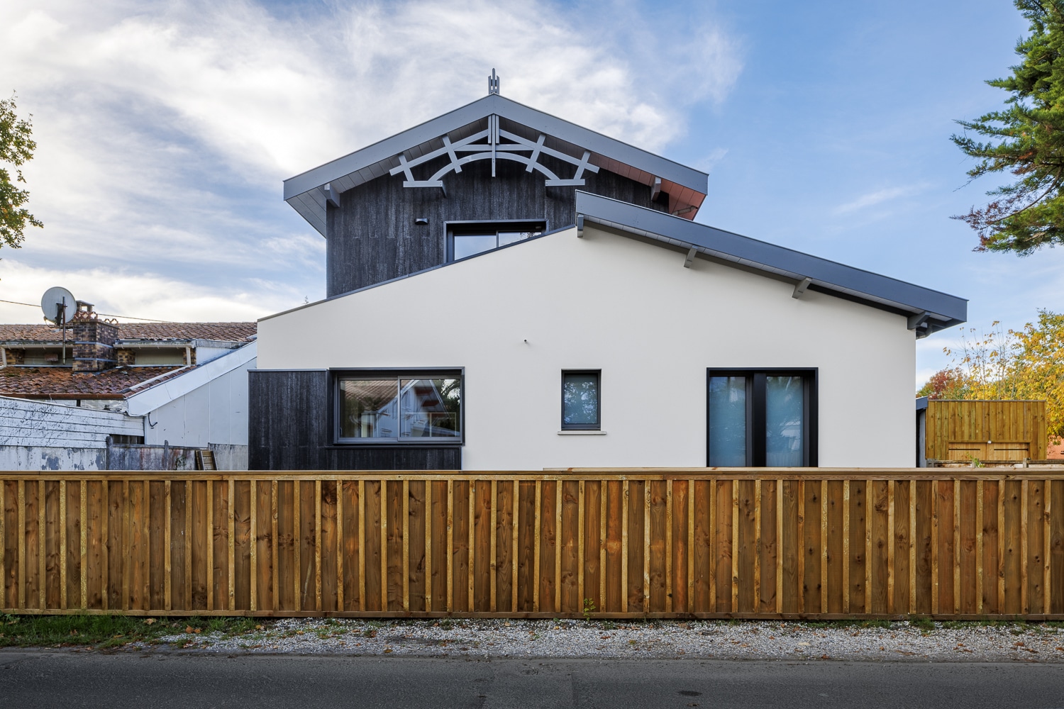 Maison contemporaine blanche et bois noir avec toiture grise, clôture en bois brun devant un ciel bleu nuageux.
