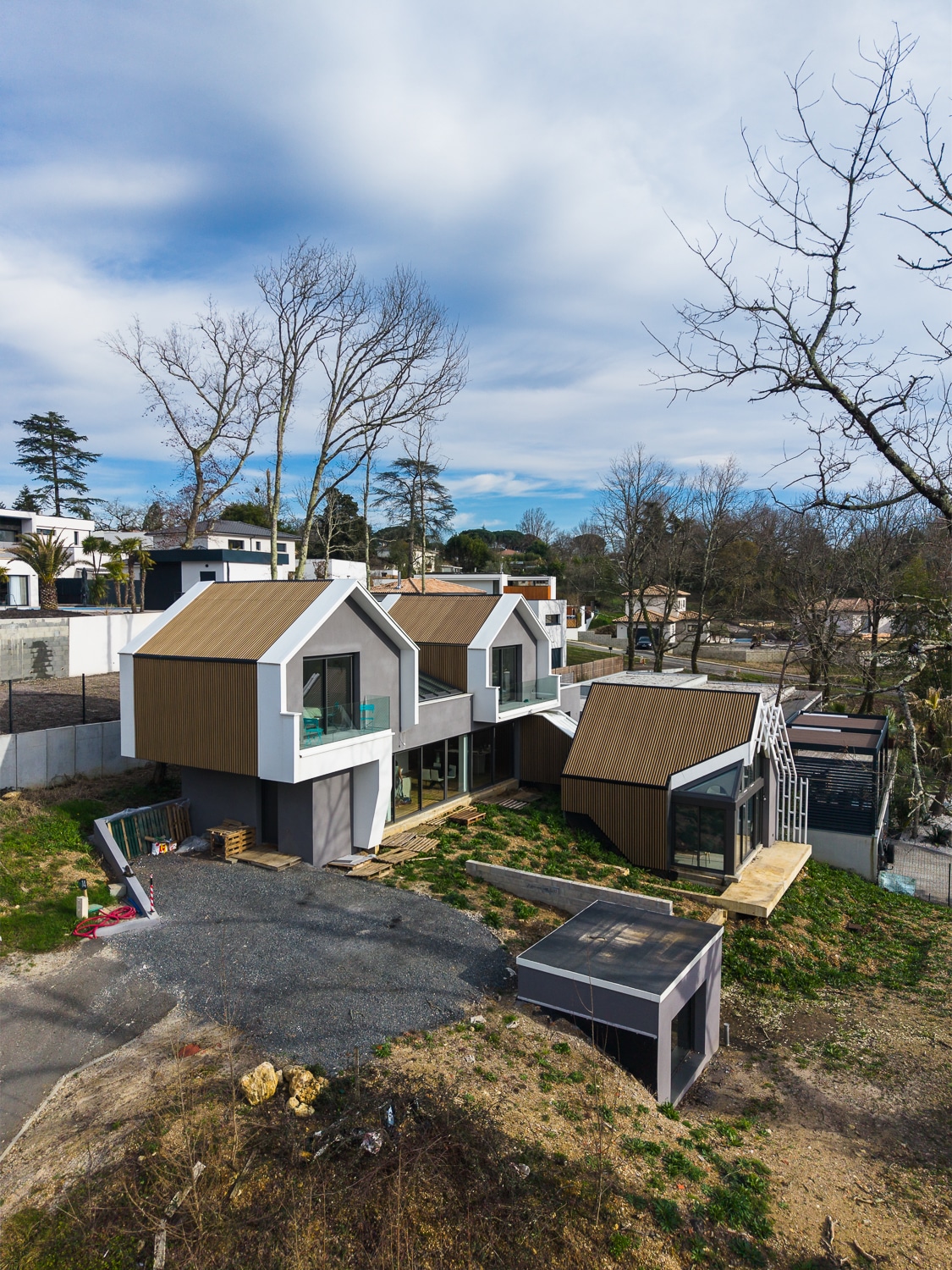 Maison d'architecte moderne aux toits pointus en bois et façades grises sur un terrain en pente sous un ciel nuageux.