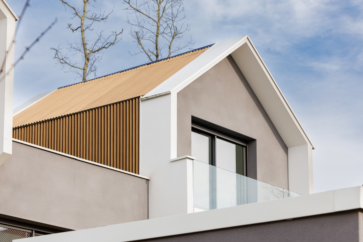 Maison moderne avec toit à pignon en bois, balcon en verre et murs gris clair sous un ciel bleu.