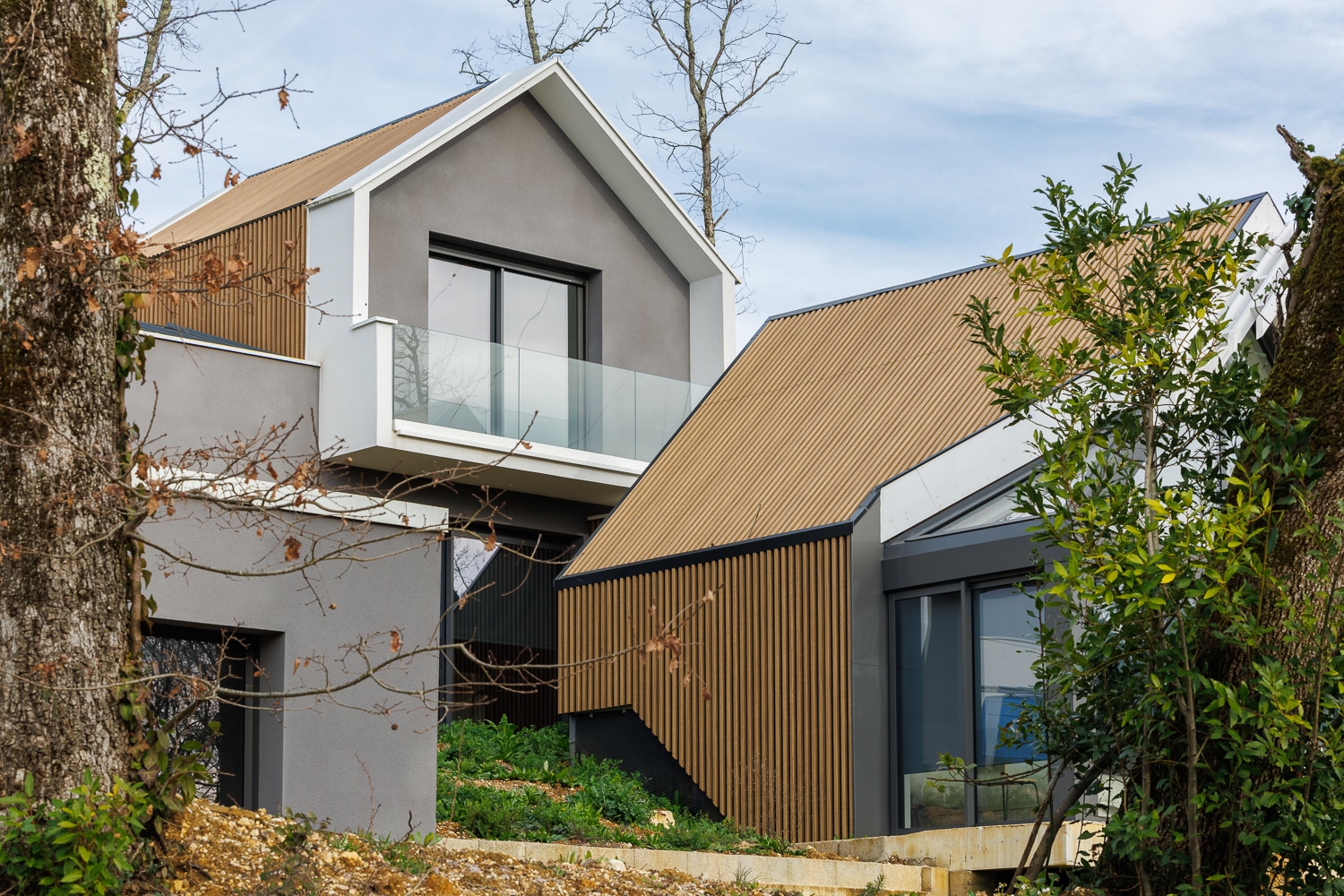 Maison moderne aux façades grises et bois avec toits en pente et grandes baies vitrées dans un cadre boisé.