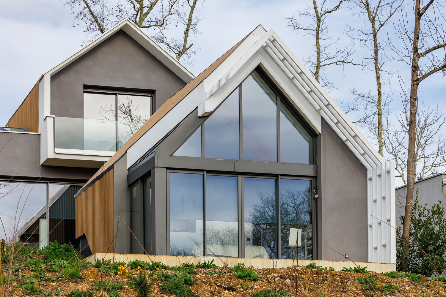 Maison moderne à toits pointus avec grandes baies vitrées, murs gris et bois dans un cadre naturel arboré.