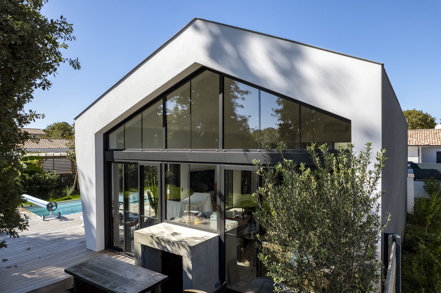 Façade blanche moderne avec grande baie vitrée triangulaire donnant sur une terrasse en bois et une piscine extérieure sous un ciel bleu clair.