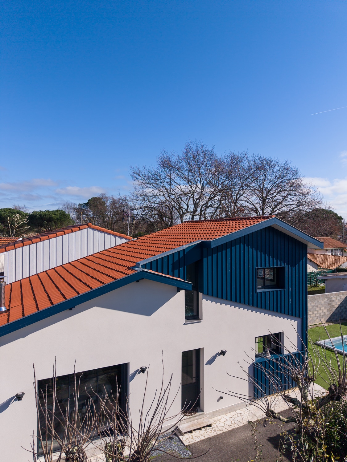 Maison contemporaine à façade blanche, bardage bleu et toiture en tuiles rouges sous un ciel dégagé.