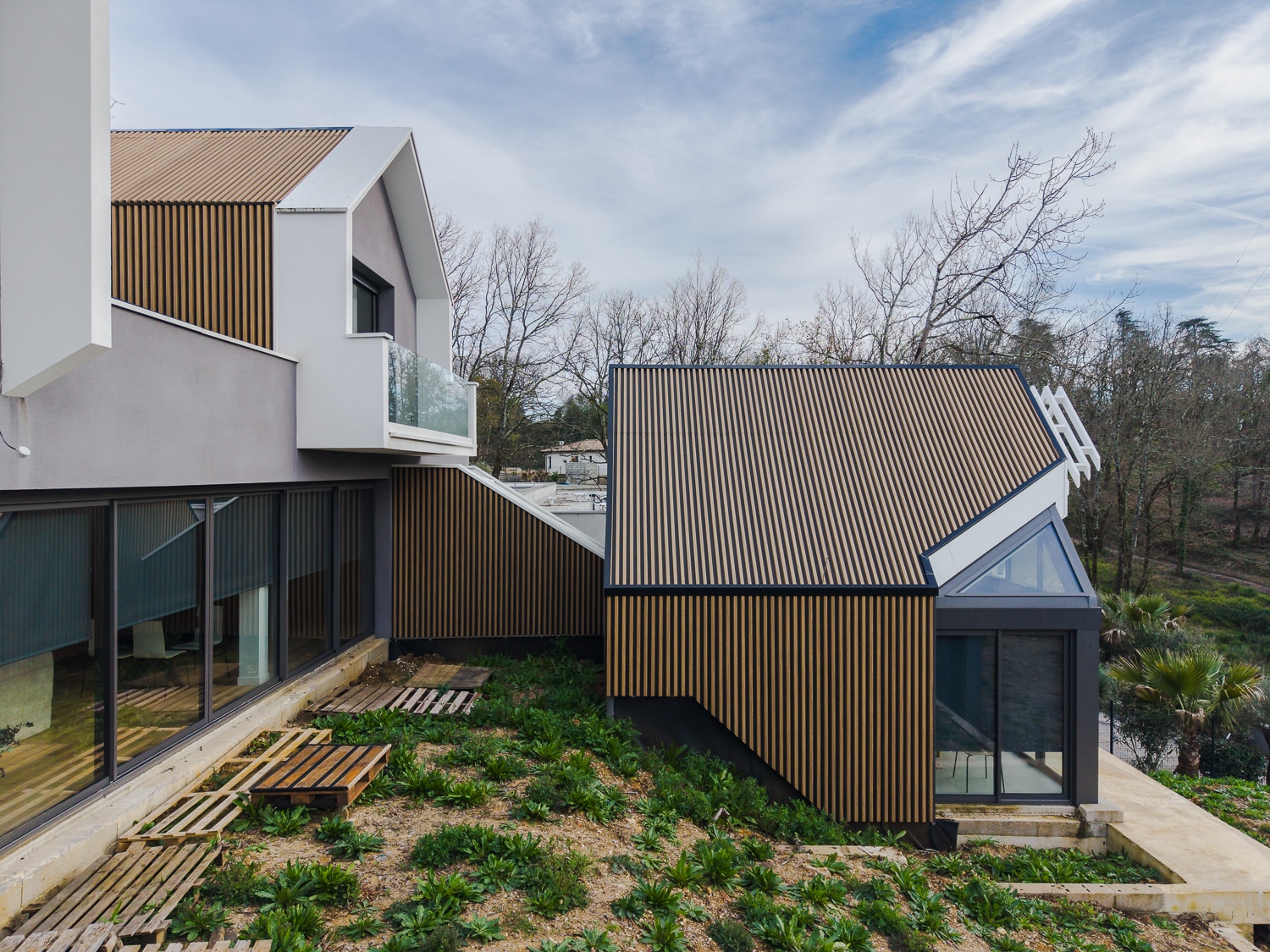 Maison moderne à bardage bois vertical et murs gris sur un terrain en pente avec de grandes baies vitrées.
