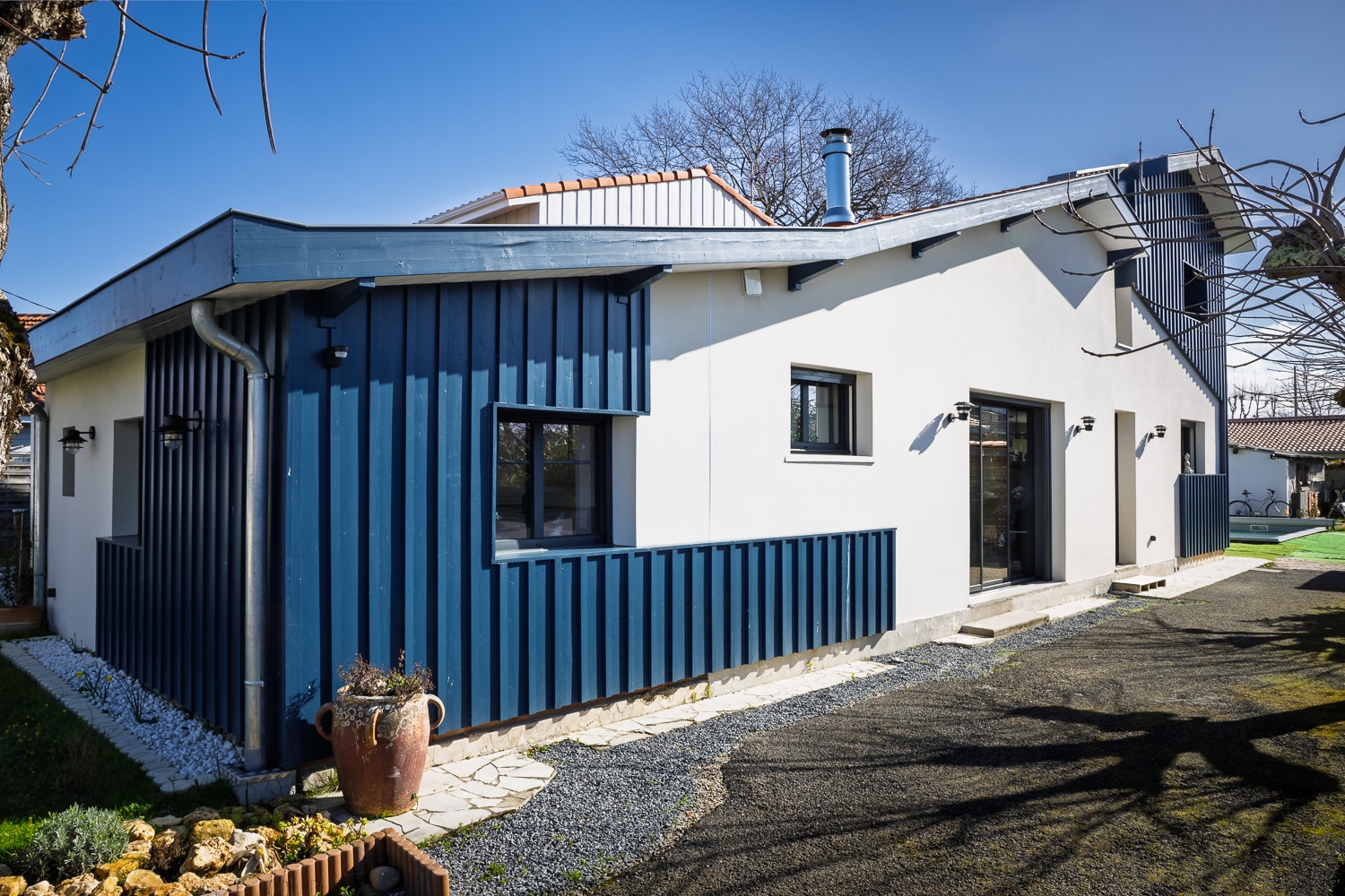 Maison contemporaine avec bardage bois bleu marine et façade blanche, allée en gravier et ciel bleu.