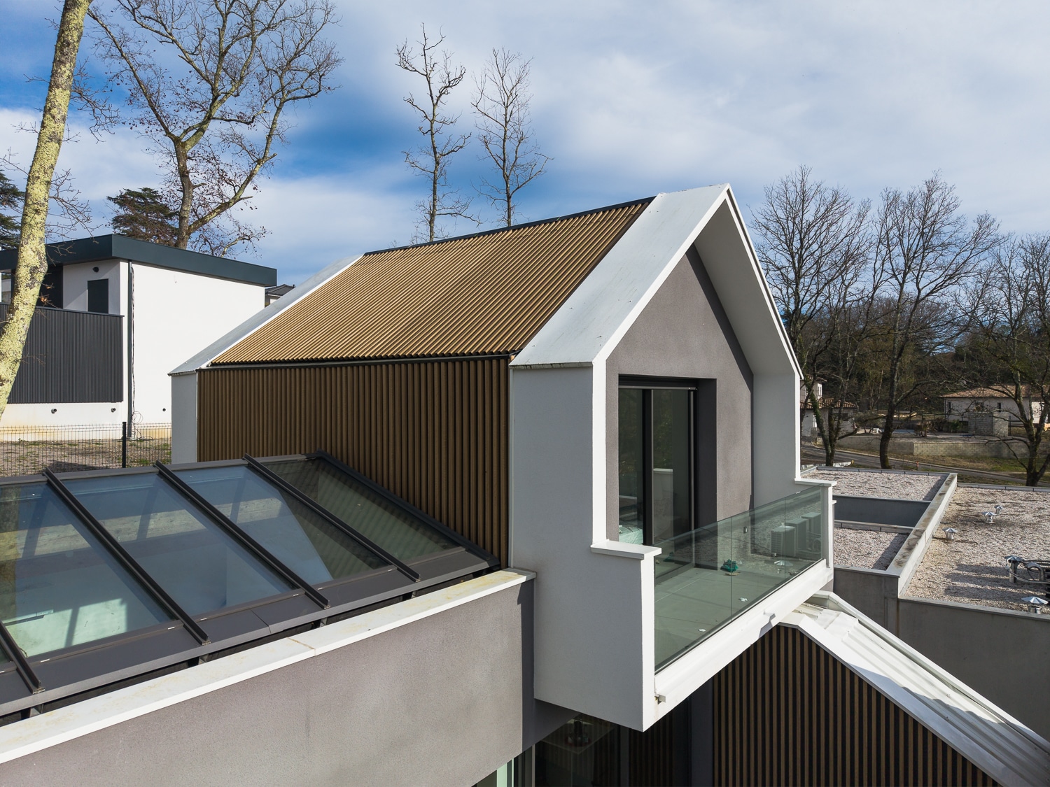 Maison moderne avec bardage bois, balcon vitré et verrière de toit sous un ciel bleu dégagé.