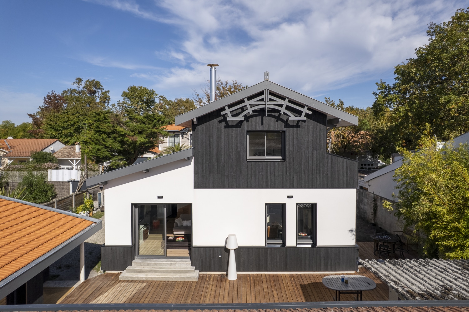 Façade d'une maison moderne mêlant enduit blanc, bardage noir et terrasse en bois, sous un ciel bleu parsemé de nuages.