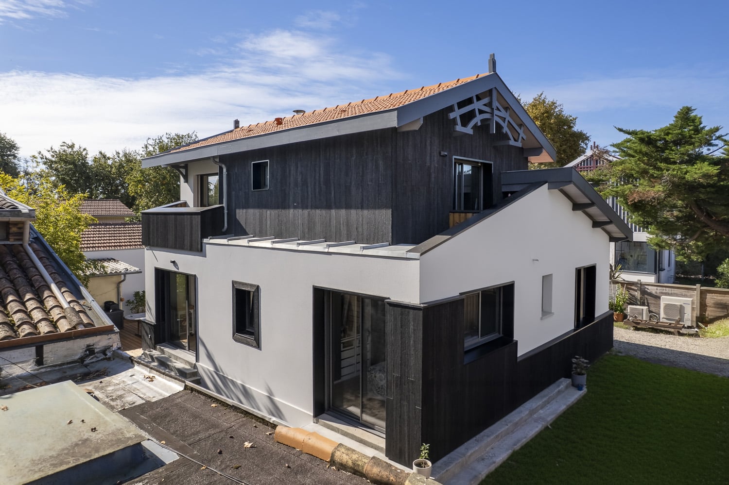 Maison moderne juxtaposant bardage bois noir et enduit blanc, toit en tuiles rouges sous un ciel bleu.