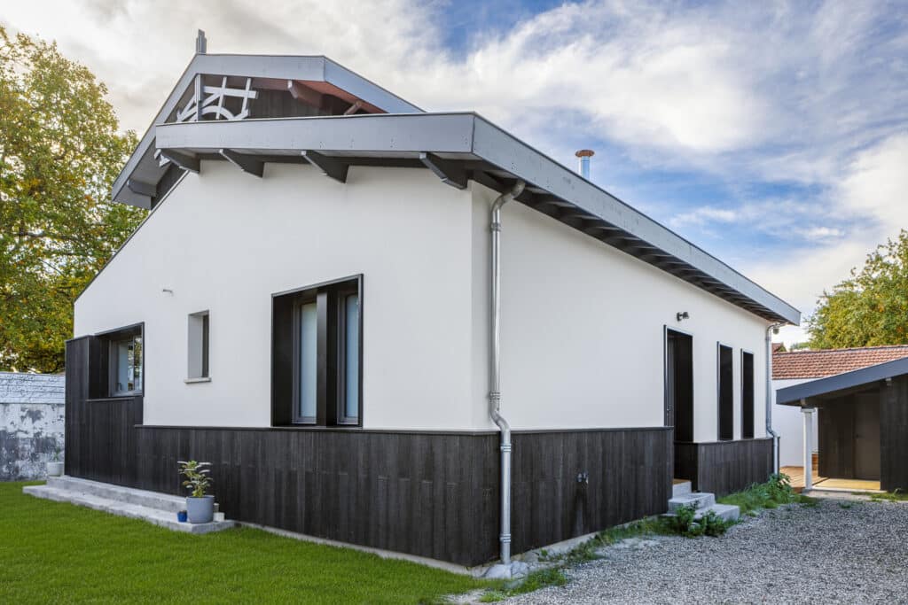 Maison moderne blanche et bois sombre avec toit à faible pente, gouttière métallique et environnement verdoyant sous un ciel nuageux.